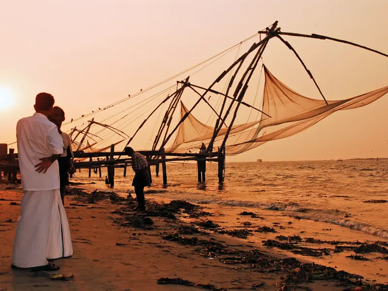 Männer in weißer Kleidung betrachten die großen Chinesischen Fischernetze am Strand von Kochi bei malerischem Sonnenuntergang.