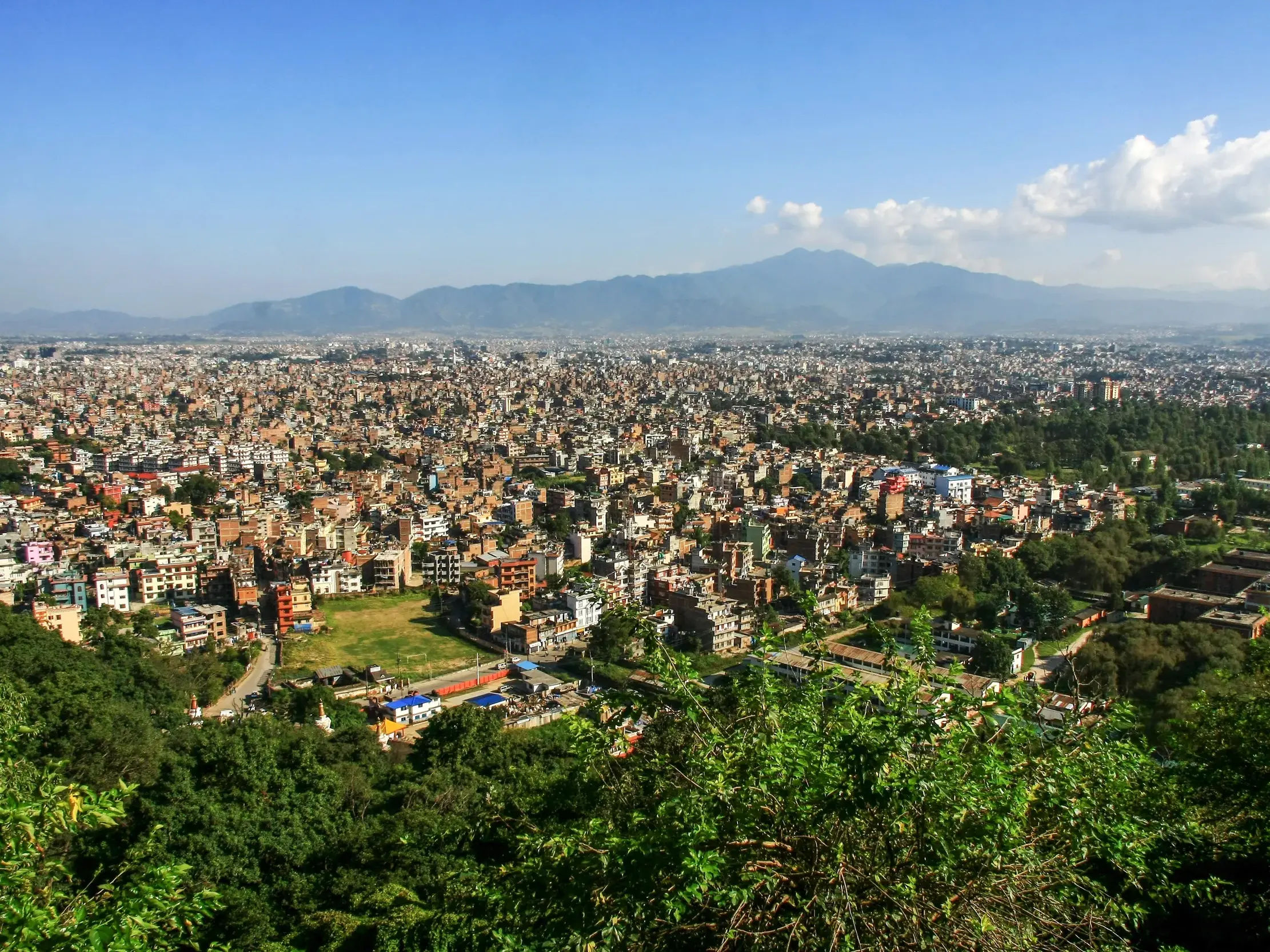 Panoramablick von einem grünen Hügel auf das weitläufige Häusermeer von Kathmandu, umgeben von der Bergkette des Tals.