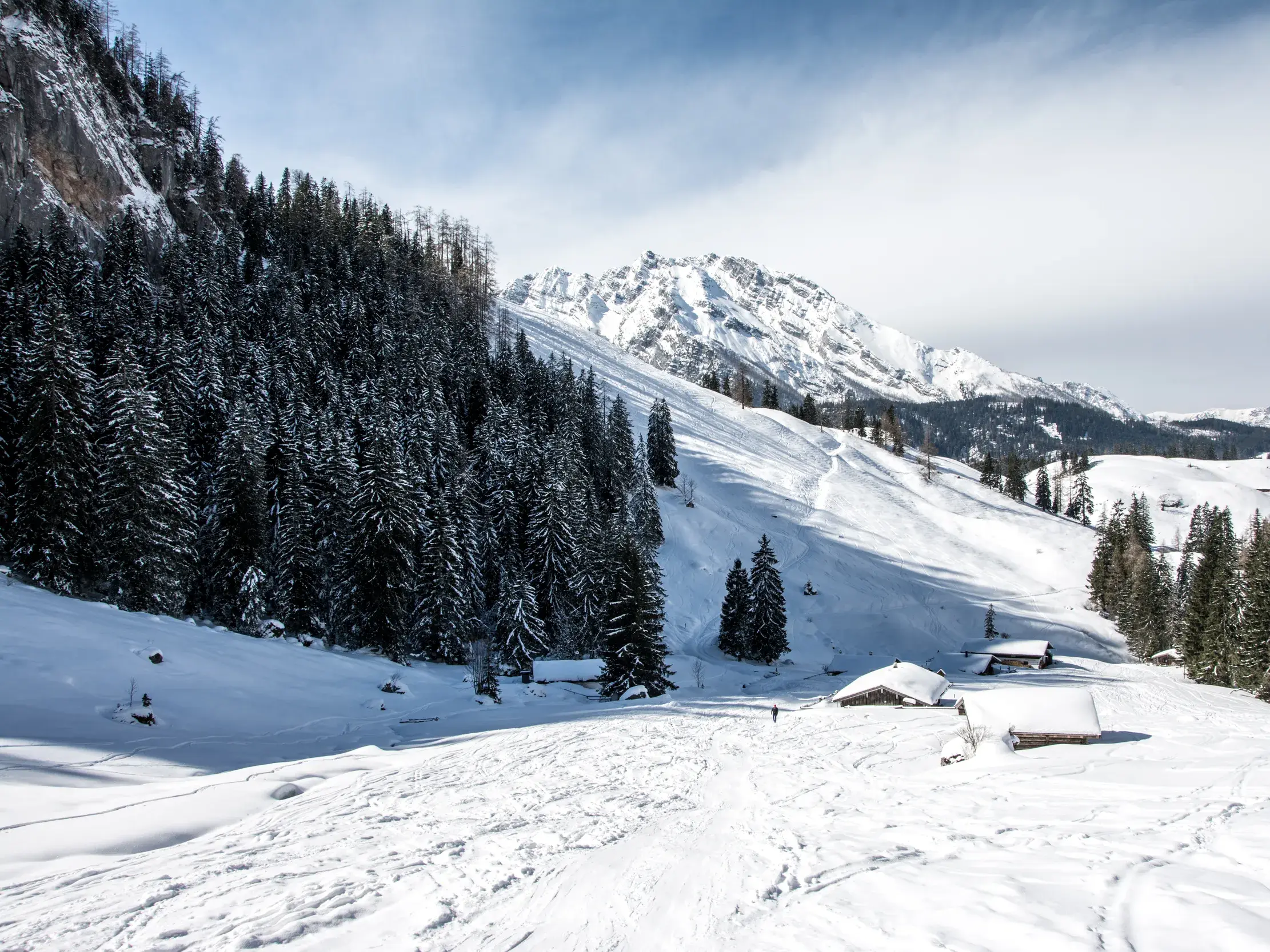 Verschneite Almhütten und Tannenwälder im Nationalpark Berchtesgaden vor der sonnigen Kulisse der Berchtesgadener Alpen.