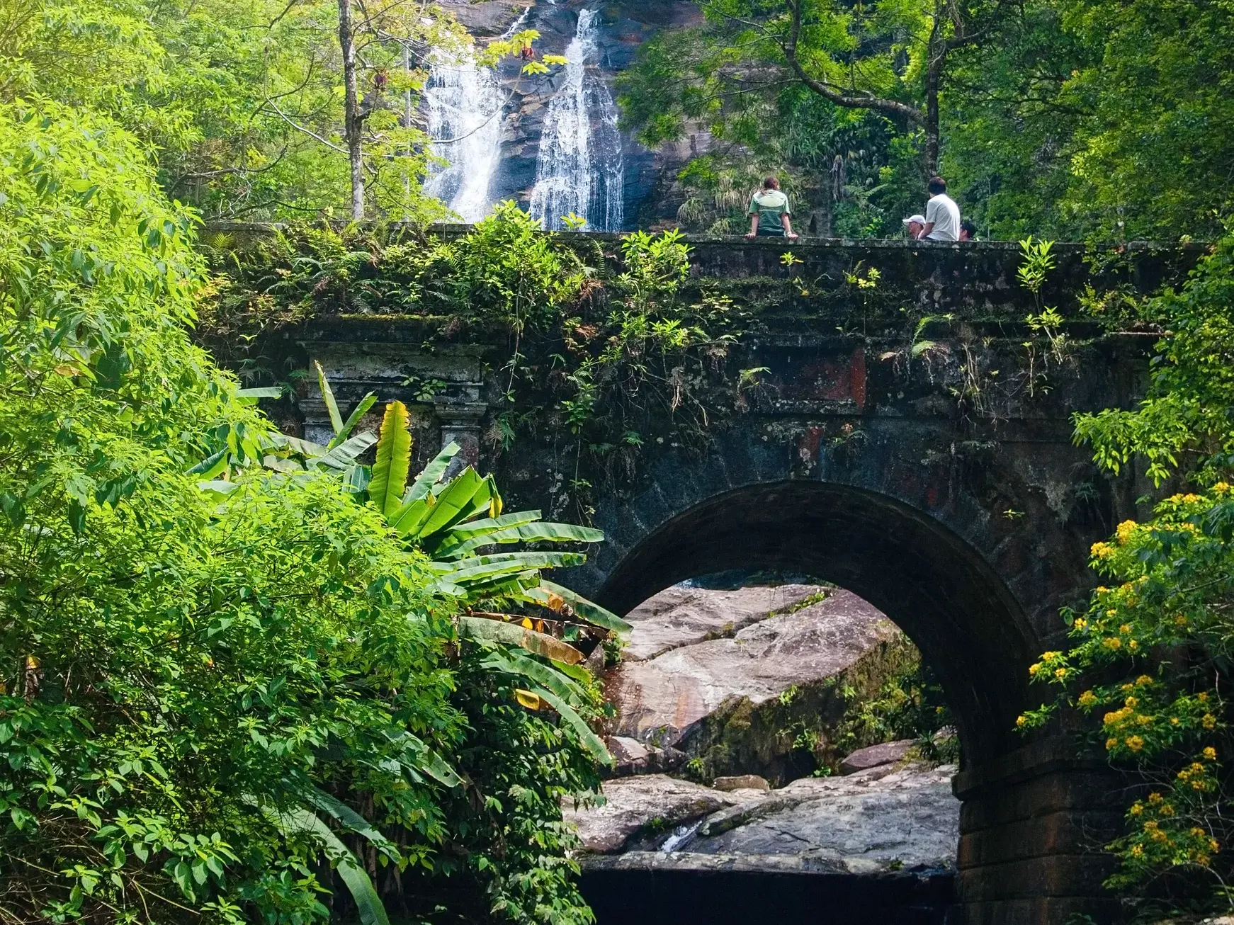 Junge Leute einer Gruppenreise entspannen auf einer alten Steinbrücke im Tijuca-Nationalpark mit Wasserfall im Hintergrund.