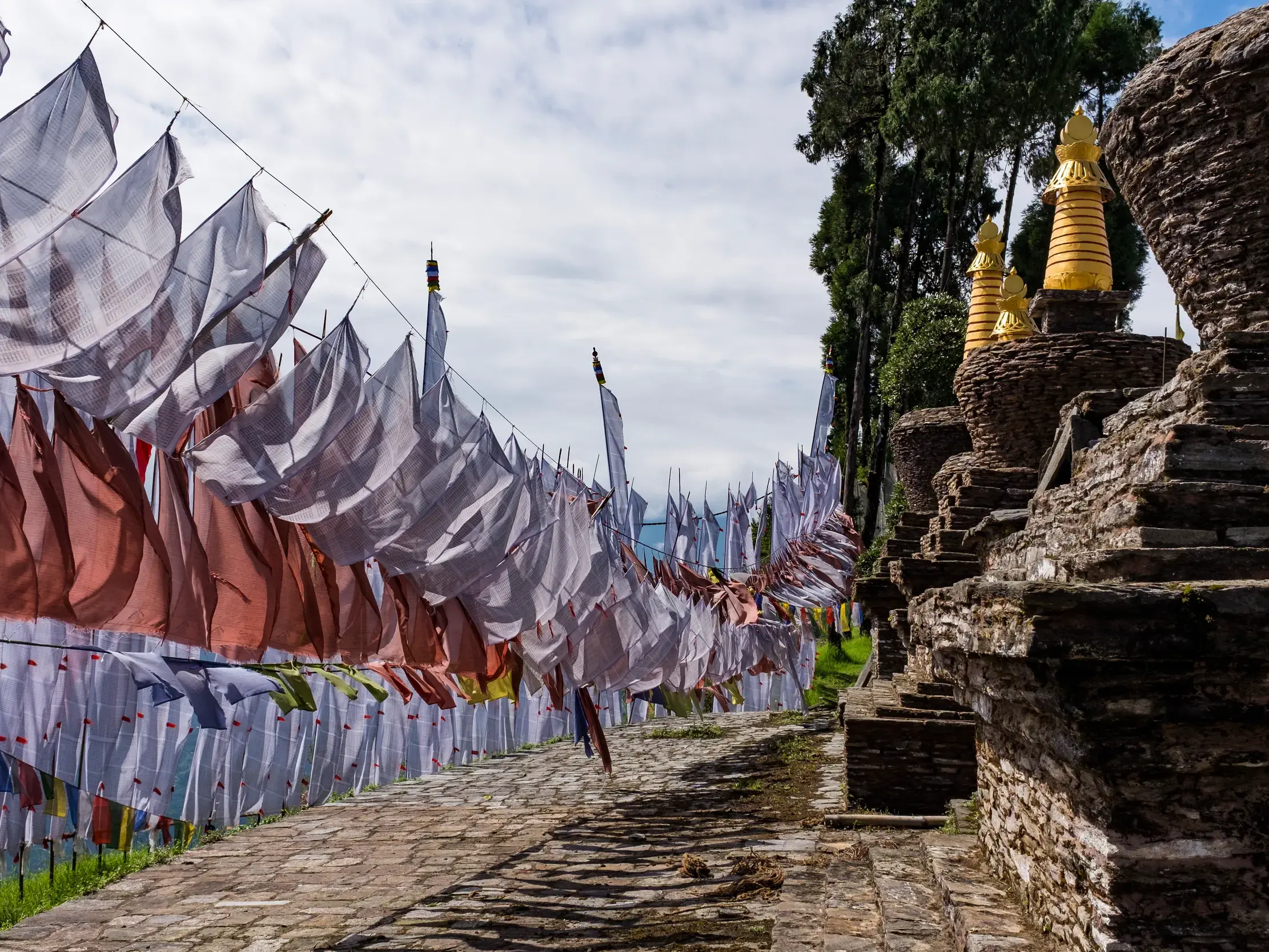 Wehende Gebetsfahnen säumen einen steinernen Trekkingpfad, der an buddhistischen Stupas mit goldenen Spitzen vorbeiführt.