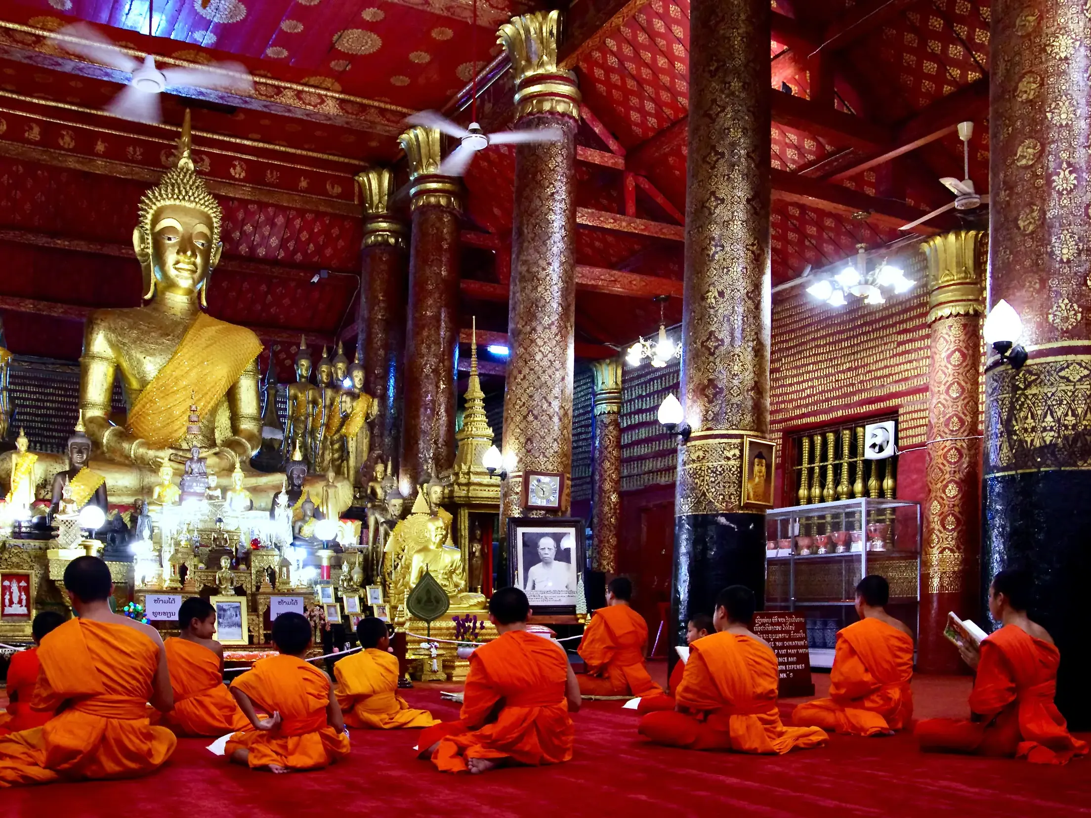 Buddhistische Mönche in orangen Roben studieren in einem prachtvollen, rot-goldenen Tempel in Laos vor einer großen Buddha-Statue.