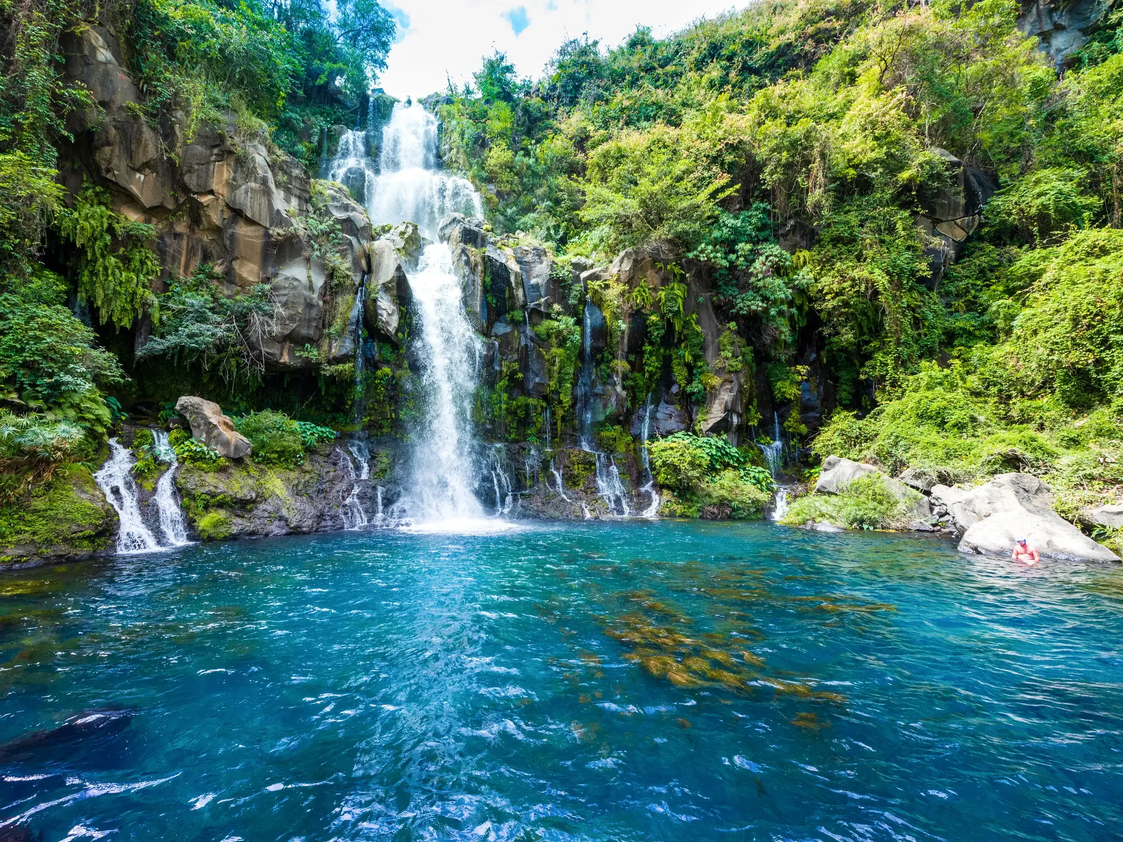 Ein Wasserfall stürzt in einen türkisen Naturpool im Dschungel von La Réunion, der auf einer Rundreise zum Baden einlädt.