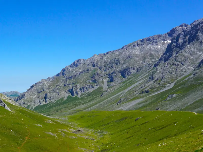 Ein grünes Gletschertal mit Wanderweg führt zu den zerklüfteten Kalksteingipfeln der Picos de Europa unter blauem Himmel.