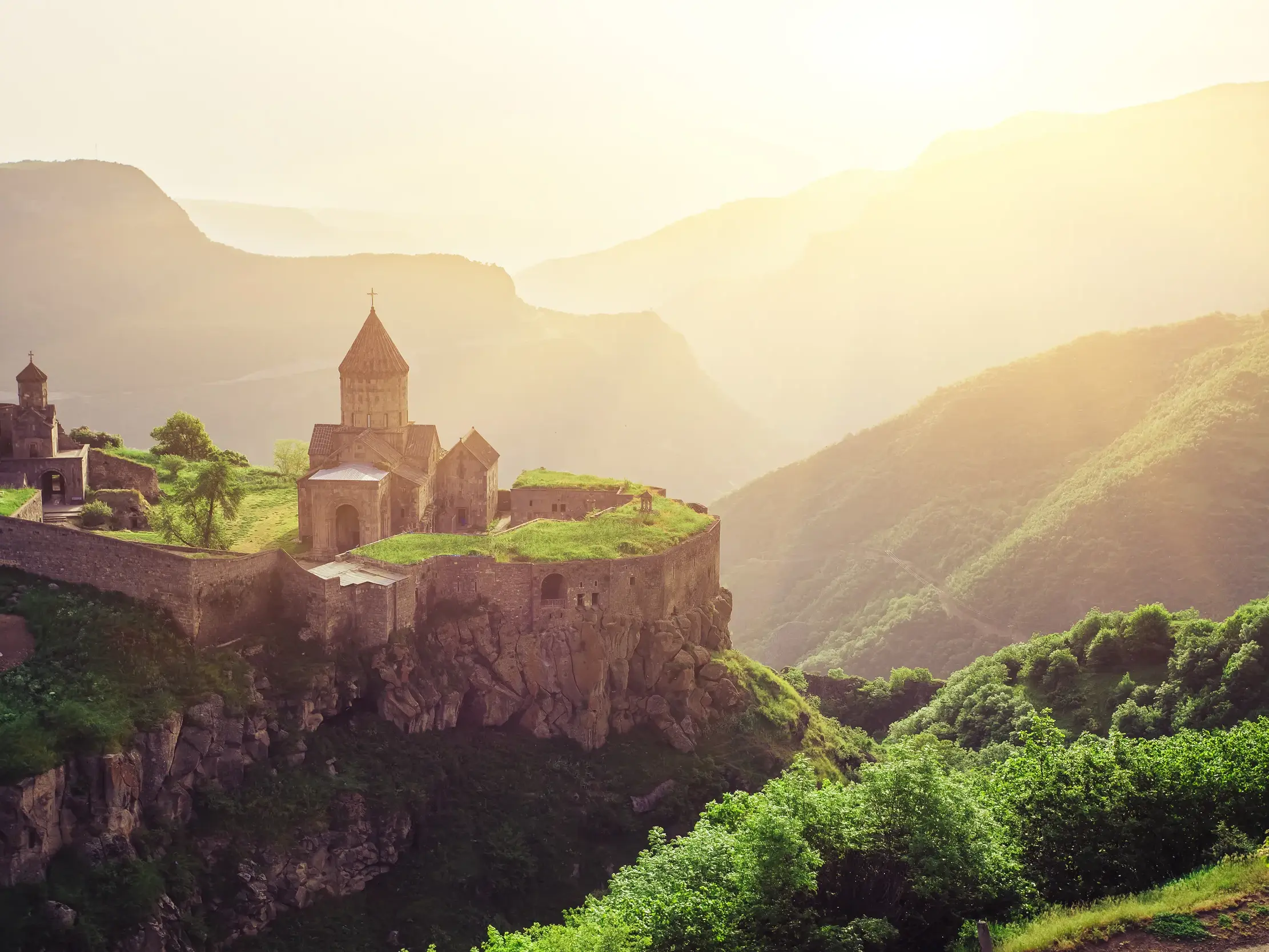 Das Kloster Tatev thront auf einem Felsen über der grünen Vorotan-Schlucht in Armenien, beleuchtet von warmem Morgenlicht.