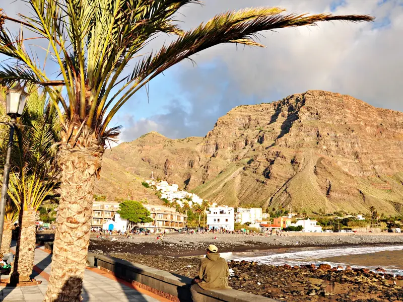 Palmen säumen die Promenade von Valle Gran Rey vor der beeindruckenden Felskulisse der Wanderregion La Gomera bei Sonnenschein.