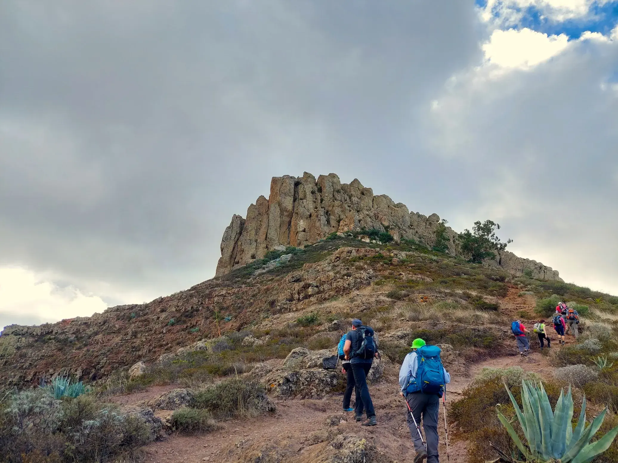 Eine Wandergruppe mit Rucksäcken und Stöcken steigt auf einem steinigen Pfad zum Tafelberg Fortaleza auf La Gomera hinauf.