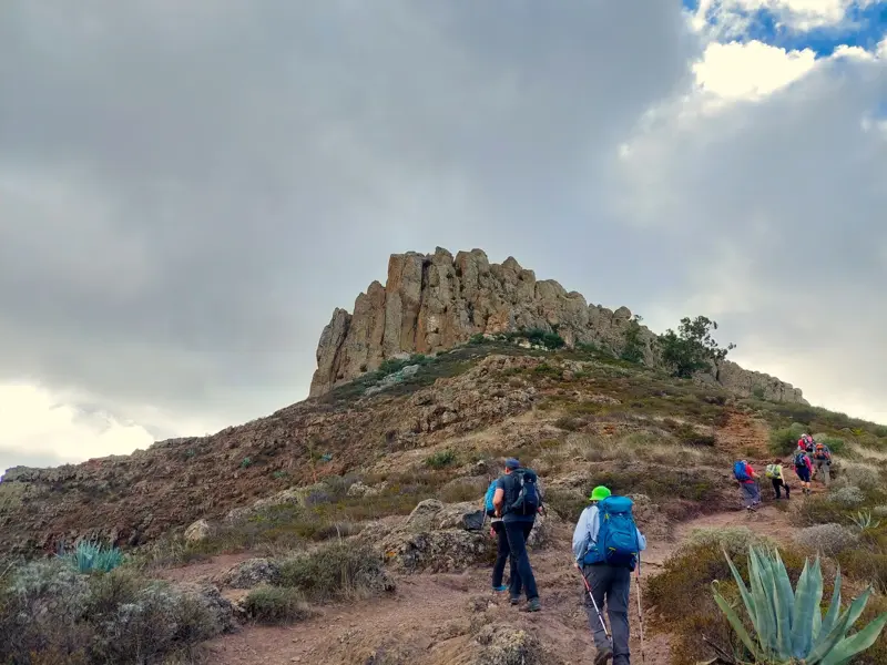 Eine Wandergruppe mit Rucksäcken und Stöcken steigt auf einem steinigen Pfad zum Tafelberg Fortaleza auf La Gomera hinauf.