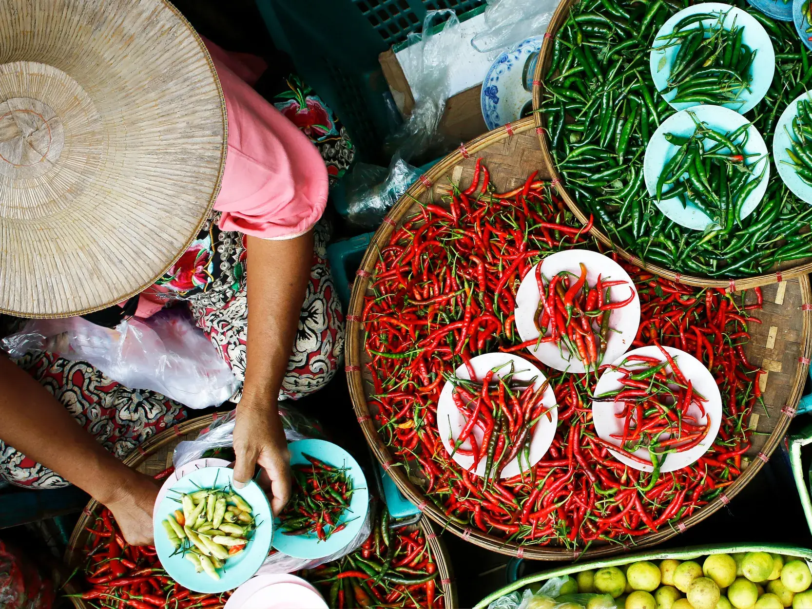 Eine Verkäuferin mit traditionellem Kegelhut sortiert auf einem Markt in Laos rote und grüne Chilischoten in Weidenkörben.