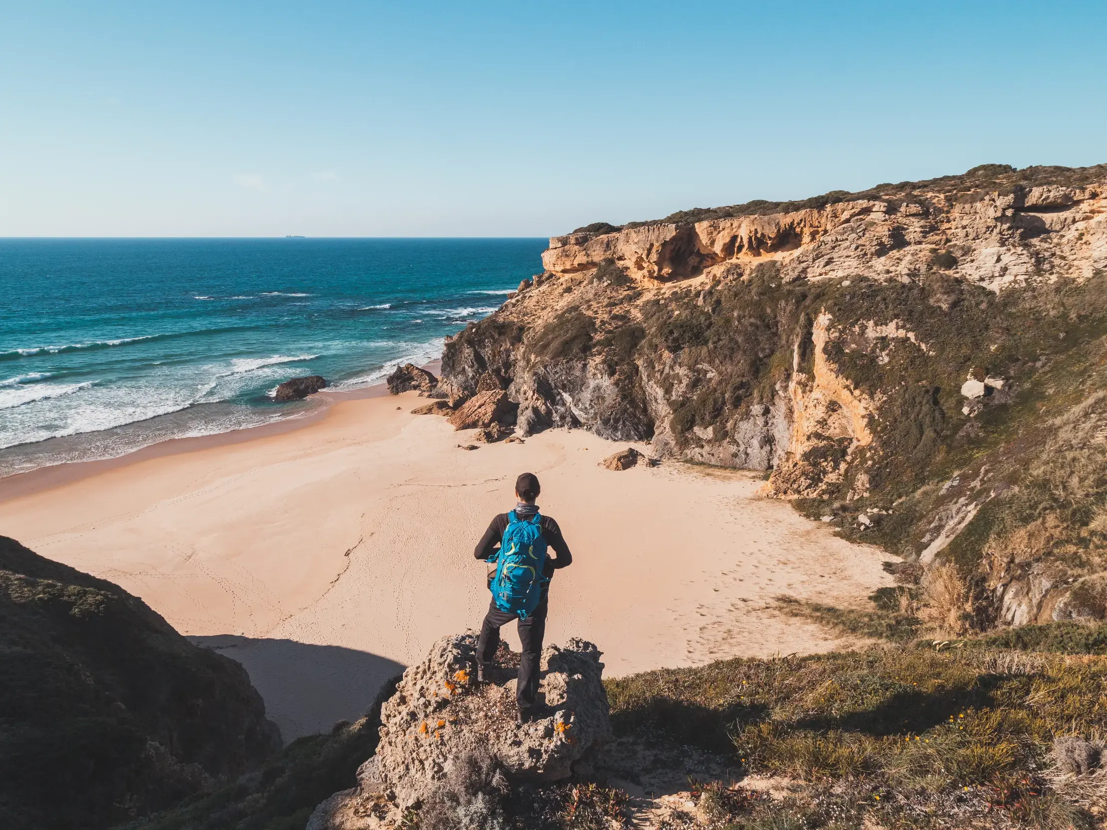 Wanderer mit Rucksack genießt auf einer Klippe den Ausblick auf den Atlantik und eine einsame Bucht am Fischerpfad der Algarve.