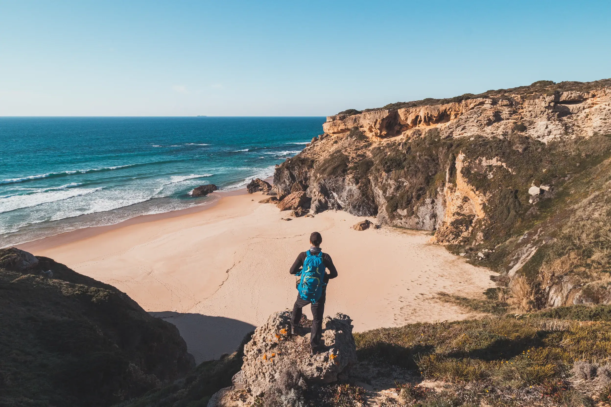 Wanderer mit Rucksack genießt auf einer Klippe den Ausblick auf den Atlantik und eine einsame Bucht am Fischerpfad der Algarve.