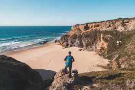 Wanderer mit Rucksack genießt auf einer Klippe den Ausblick auf den Atlantik und eine einsame Bucht am Fischerpfad der Algarve.