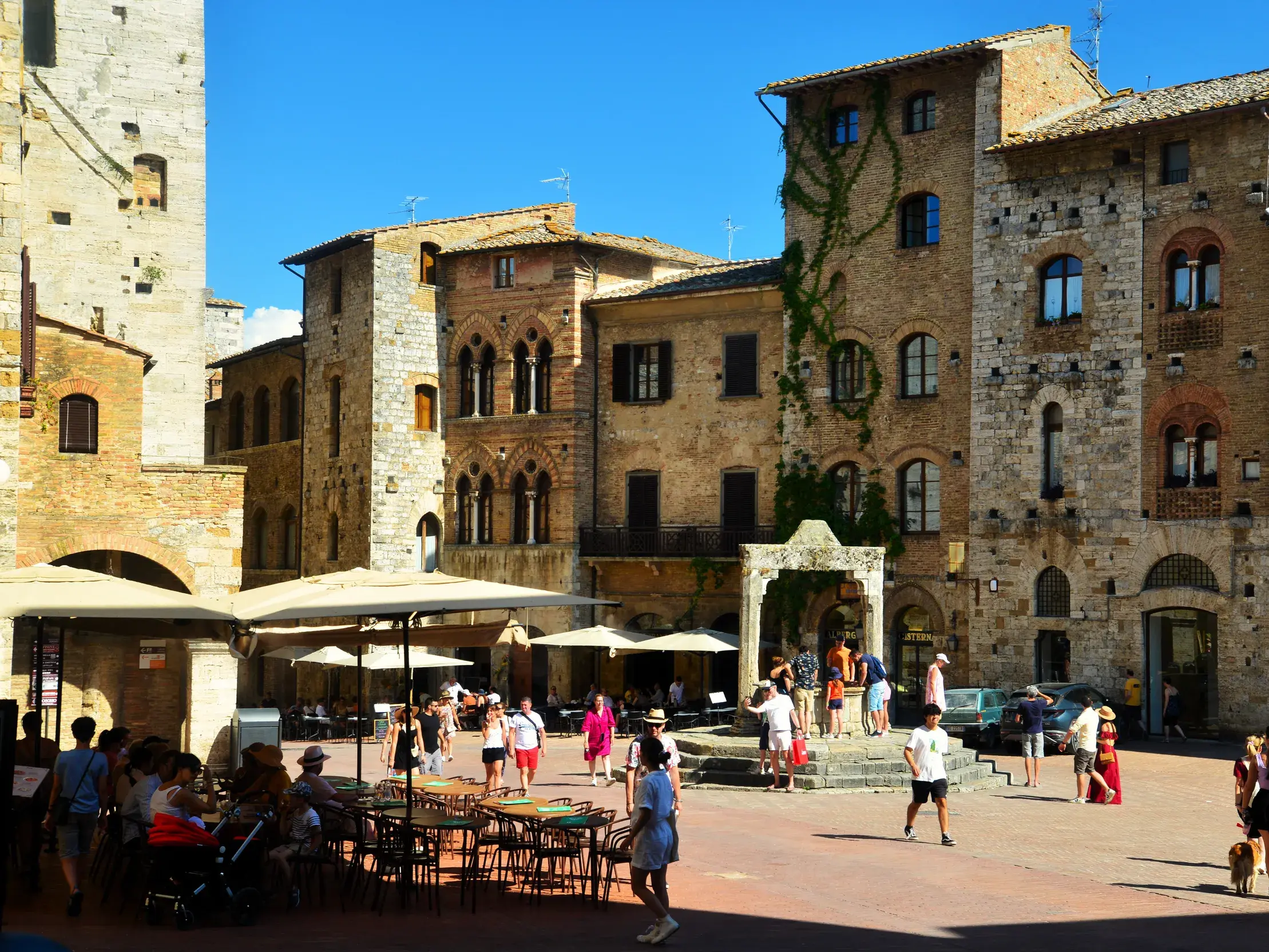 Touristen auf dem sonnigen Platz Piazza della Cisterna in San Gimignano, umgeben von mittelalterlichen Steinhäusern der Toskana.
