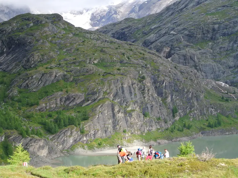Eine kleine Wandergruppe rastet auf einer Wiese am Kapruner Stausee mit Blick auf die vergletscherten Hohen Tauern.
