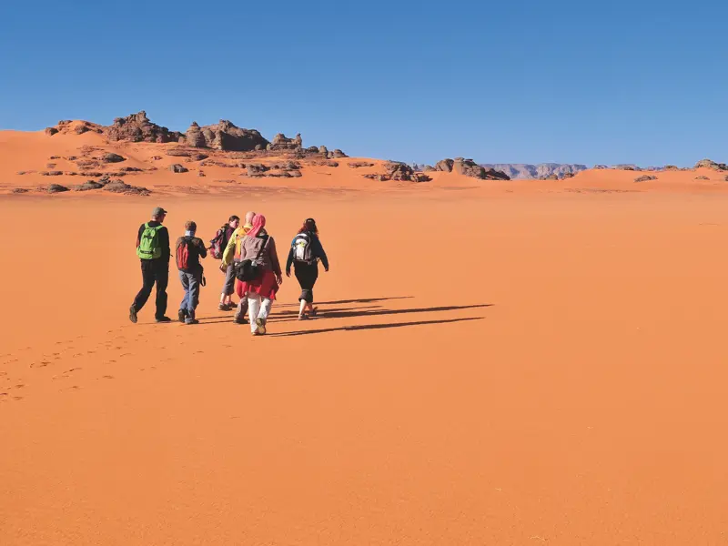 Eine kleine Reisegruppe mit Rucksäcken beim Trekking durch die weite, orange Sandwüste des Tadrart in Algerien.