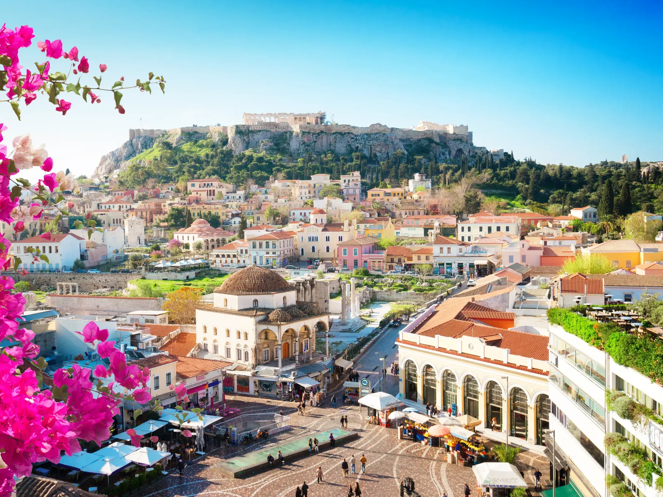 Sonniger Blick über den belebten Monastiraki-Platz in Athen, mit leuchtender Bougainvillea und der Akropolis im Hintergrund.