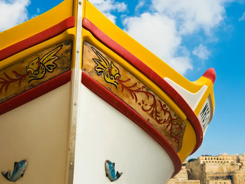 Nahaufnahme des bunten Bugs eines traditionellen Luzzu-Bootes in Malta mit Blick auf die historische Skyline von Valletta.