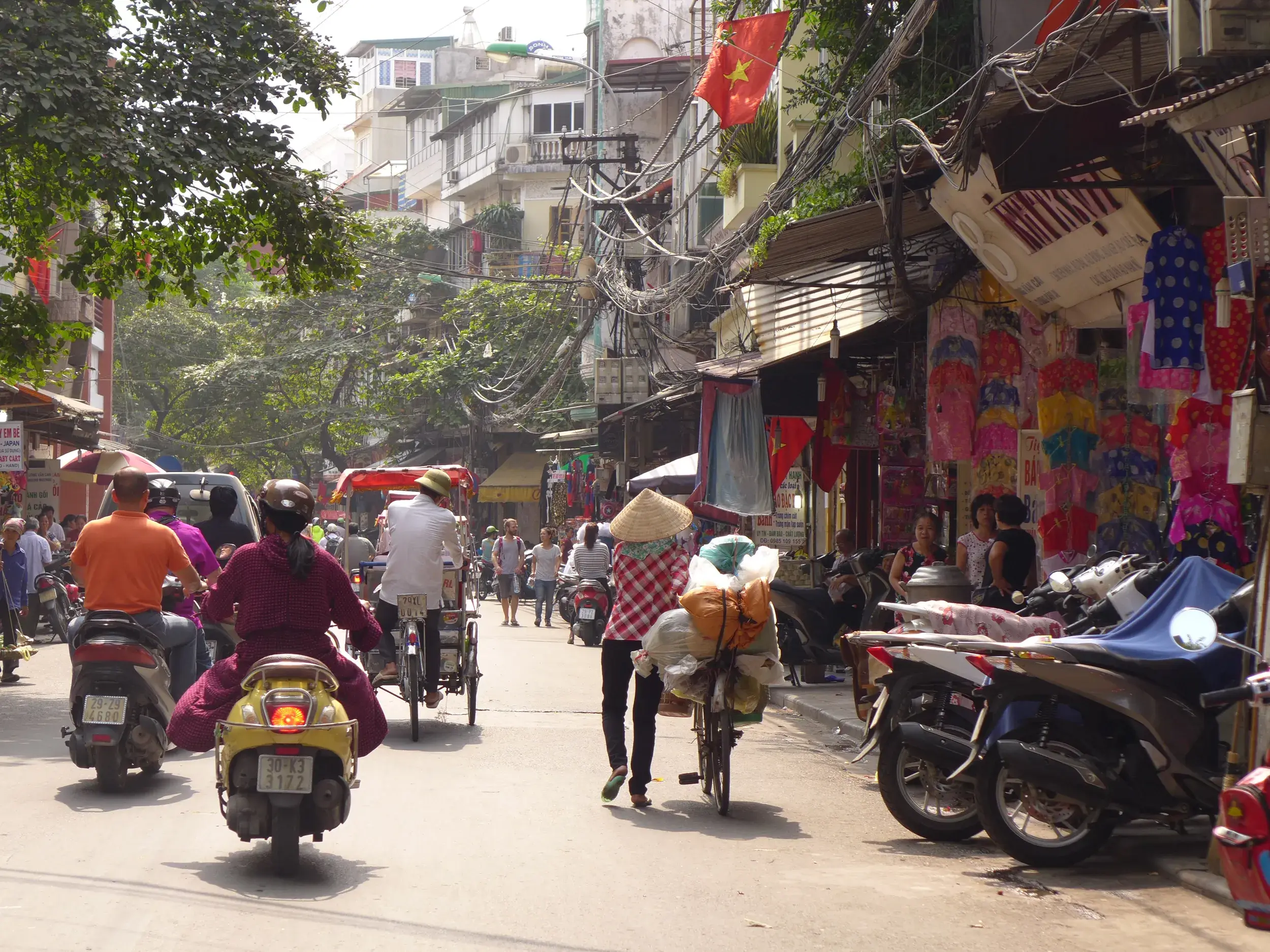 Quirliger Alltag in einer Gasse der Altstadt von Hanoi mit Mopeds, einem bepackten Fahrrad und bunten Geschäften am Straßenrand.