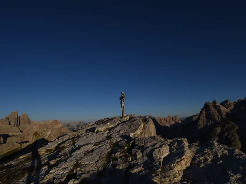 Ein Gipfelkreuz auf der felsigen Oberbachernspitze bei klarem Himmel und Sonnenaufgang über den Dolomiten.