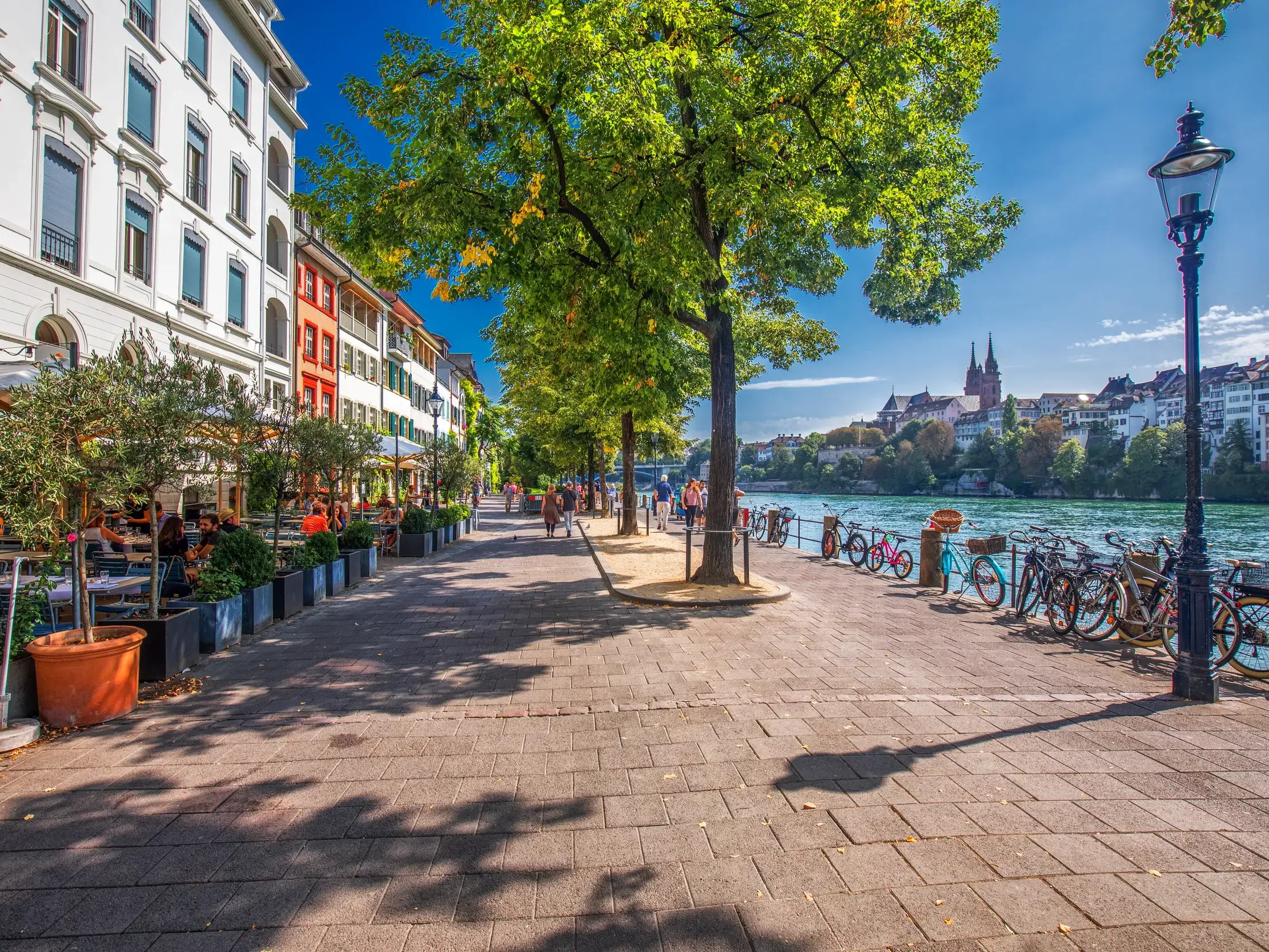 Die Rheinpromenade in Basel an einem Sonnentag mit Cafés, Spaziergängern und Blick auf das historische Basler Münster.