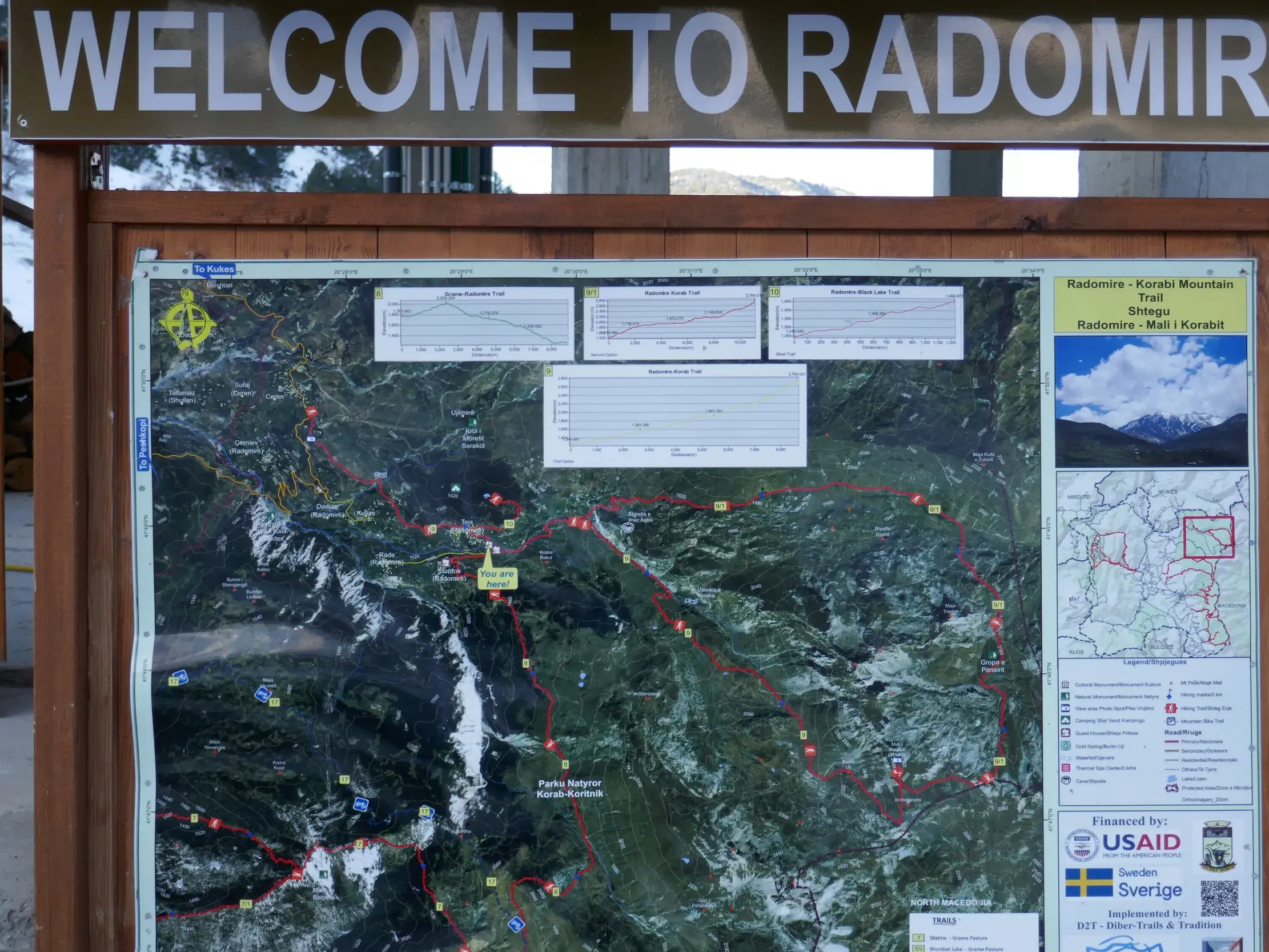 Willkommensschild in Radomire über einer Informationstafel mit Wanderkarte für Touren im Korab-Koritnik Naturpark in Albanien.