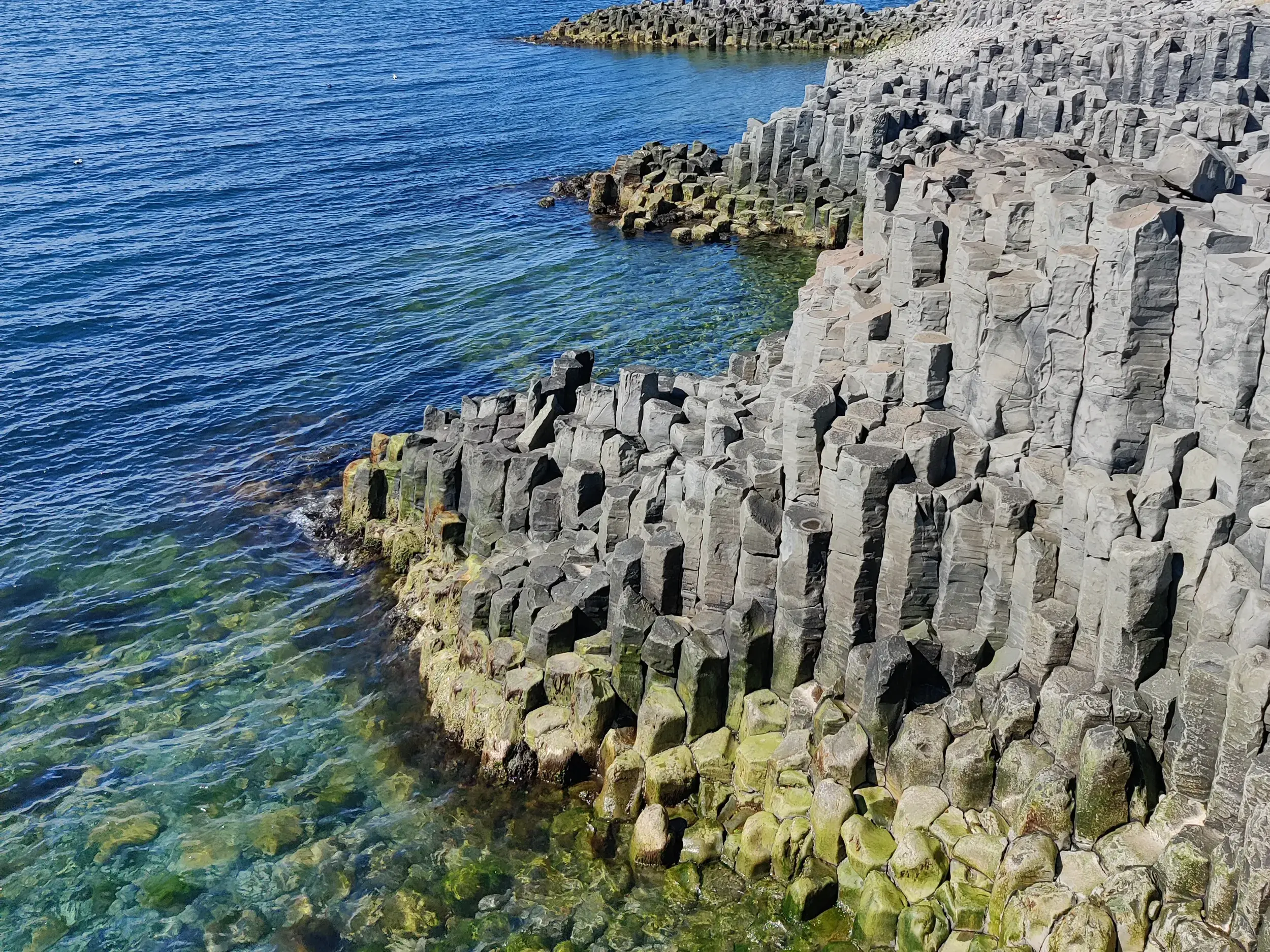 Massive, sechseckige Basaltsäulen an Islands Küste ragen bei Sonnenschein aus dem klaren, türkisfarbenen Ozean.