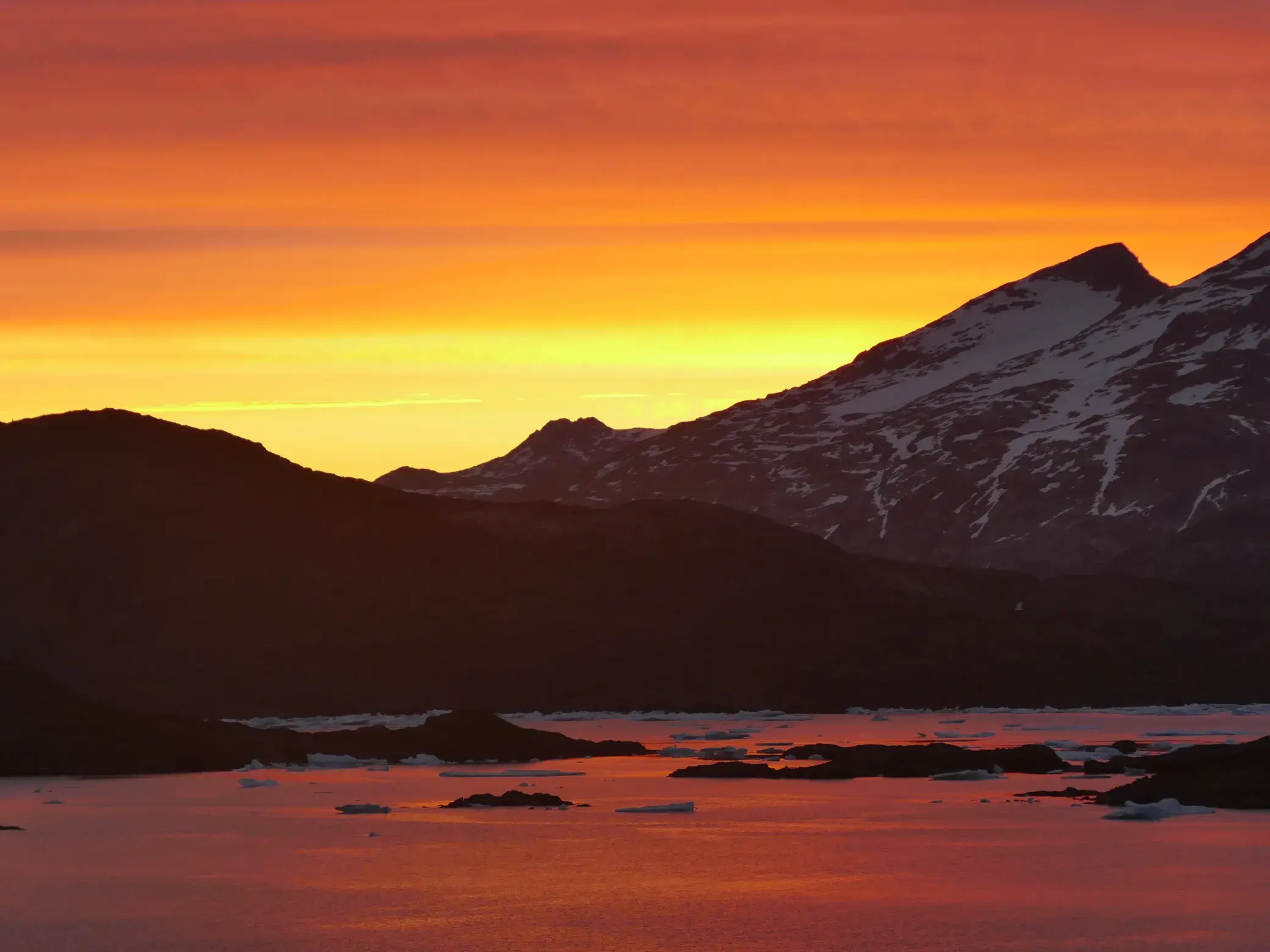 Die Mitternachtssonne taucht schneebedeckte Berge und einen Fjord mit Eisschollen in Ostgrönland in orangerotes Licht.