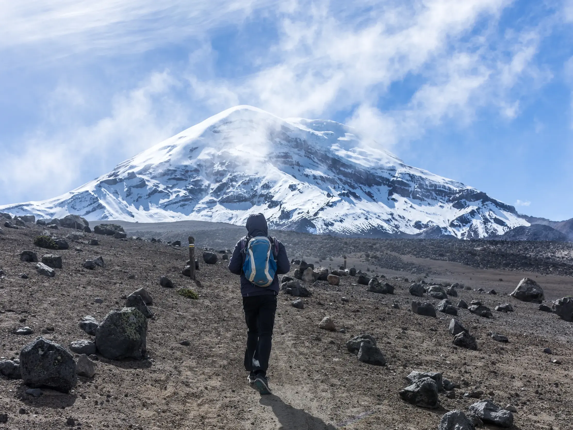 Ein Wanderer mit Rucksack geht auf einem steinigen Pfad in Ecuador mit Blick auf den schneebedeckten Gipfel des Vulkans Chimborazo.