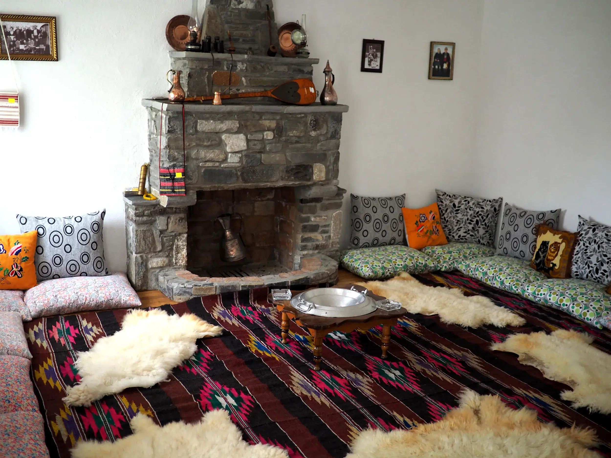 Gemütliche Sitzecke in einem albanischen Gästehaus im Korab-Gebirge mit Steinkamin, bunten Kissen und traditionellem Teppich.