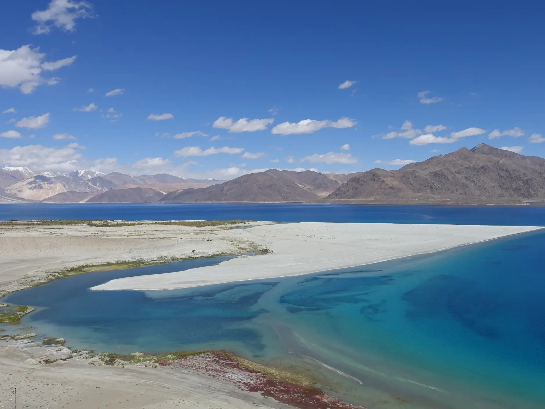 Panoramablick auf den tiefblauen Pangong See in Ladakh mit einer weißen Landzunge unter blauem Himmel und Himalaya-Gipfeln.