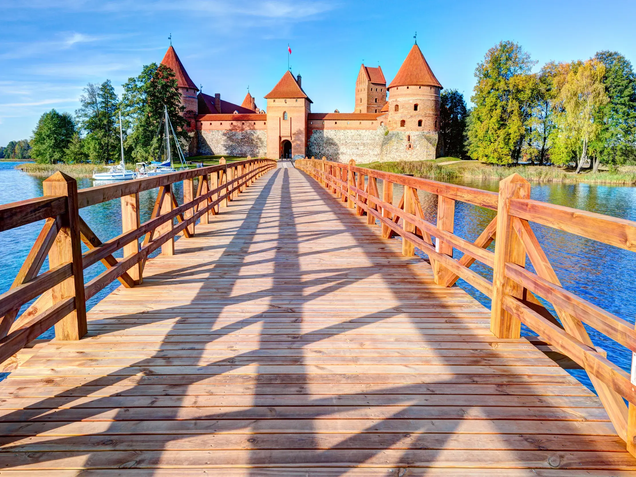 Langer Holzsteg mit Geländerschatten führt über den blauen See zur gotischen Wasserburg Trakai in Litauen an einem Sonnentag.