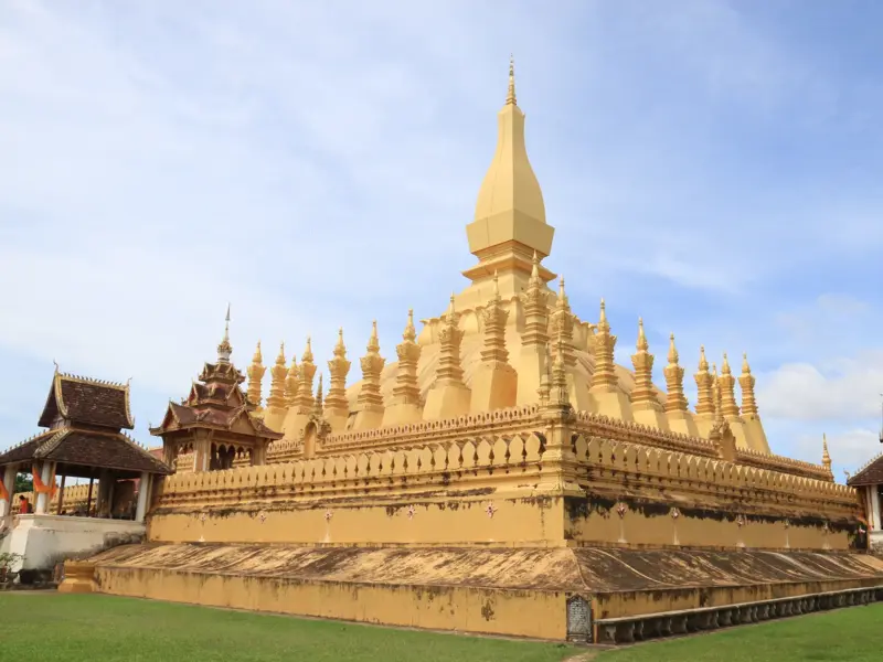 Die große, goldene Stupa Pha That Luang, das Nationalsymbol von Laos, auf einer Wiese unter blauem Himmel in Vientiane.