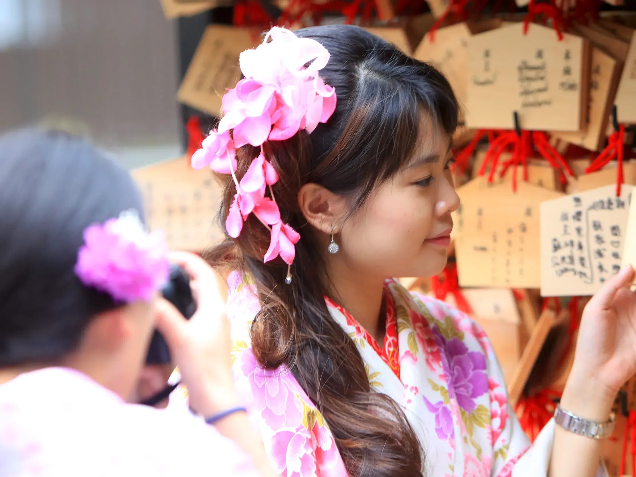 Junge Frau im Kimono mit Kirschblütenschmuck betrachtet auf einer Studienreise in Japan die Ema-Wunschtafeln an einem Schrein.