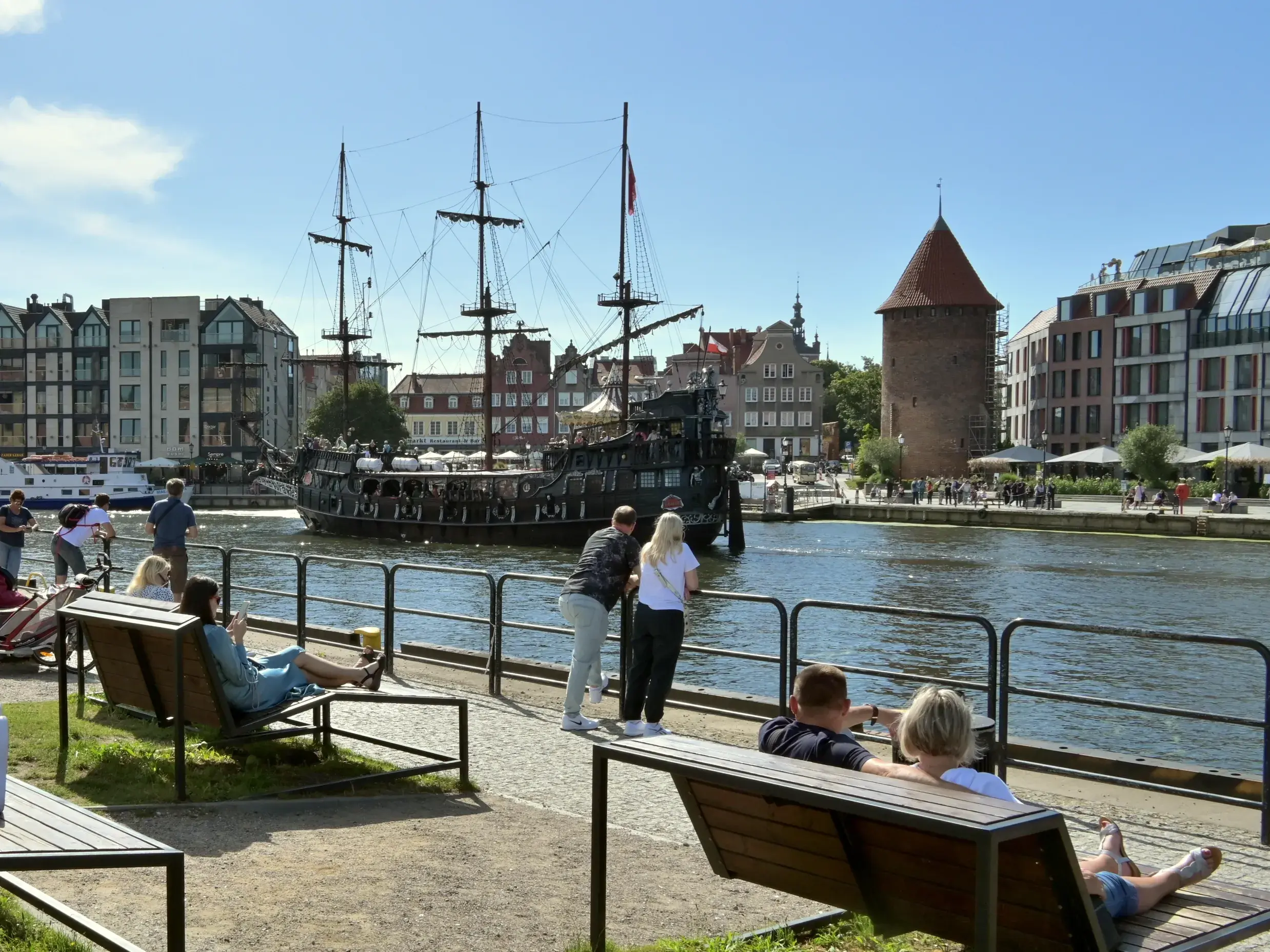 Menschen entspannen an der Uferpromenade der Mottlau in Danzig und genießen bei Sonnenschein den Blick auf ein historisches Schiff.