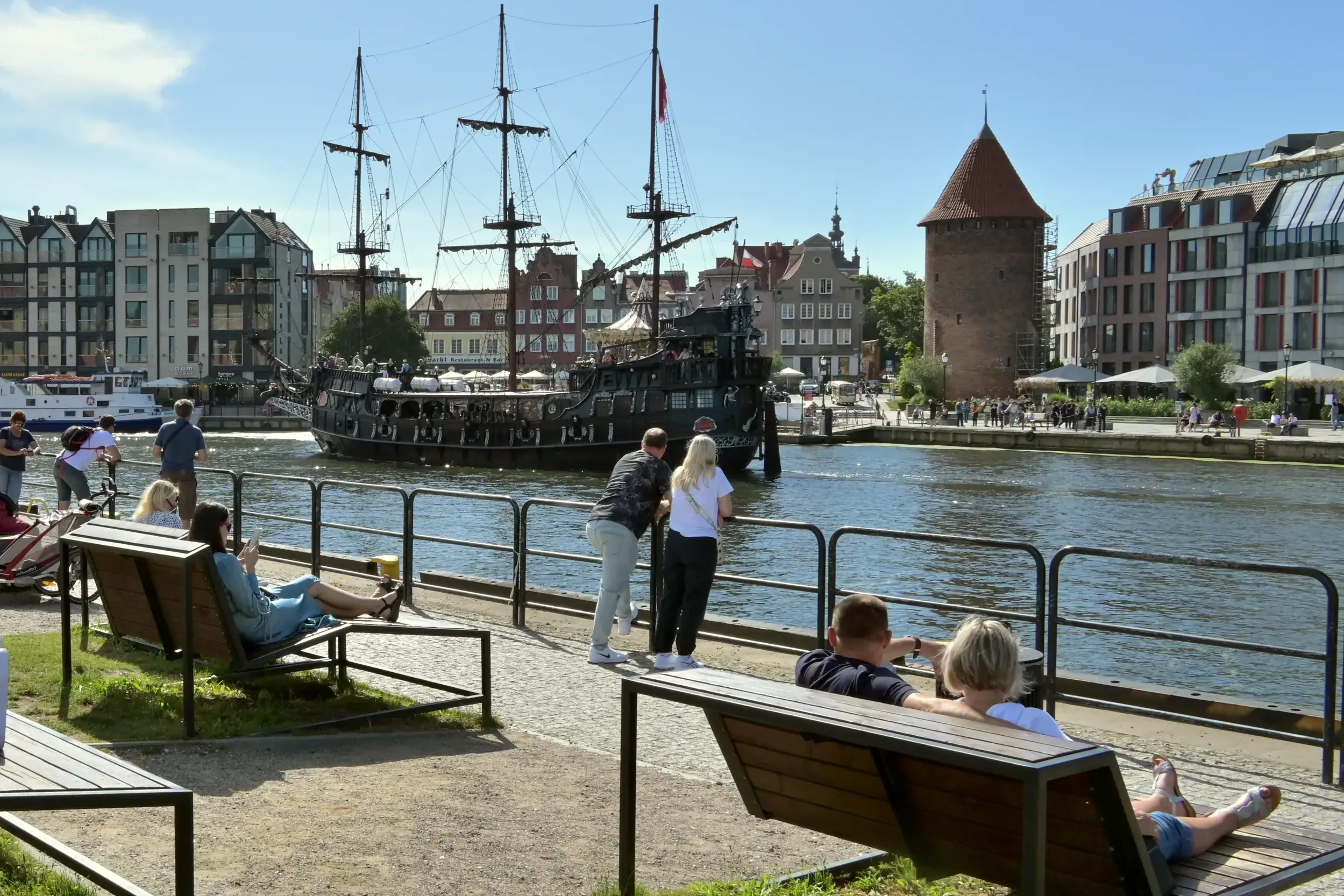 Menschen entspannen an der Uferpromenade der Mottlau in Danzig und genießen bei Sonnenschein den Blick auf ein historisches Schiff.