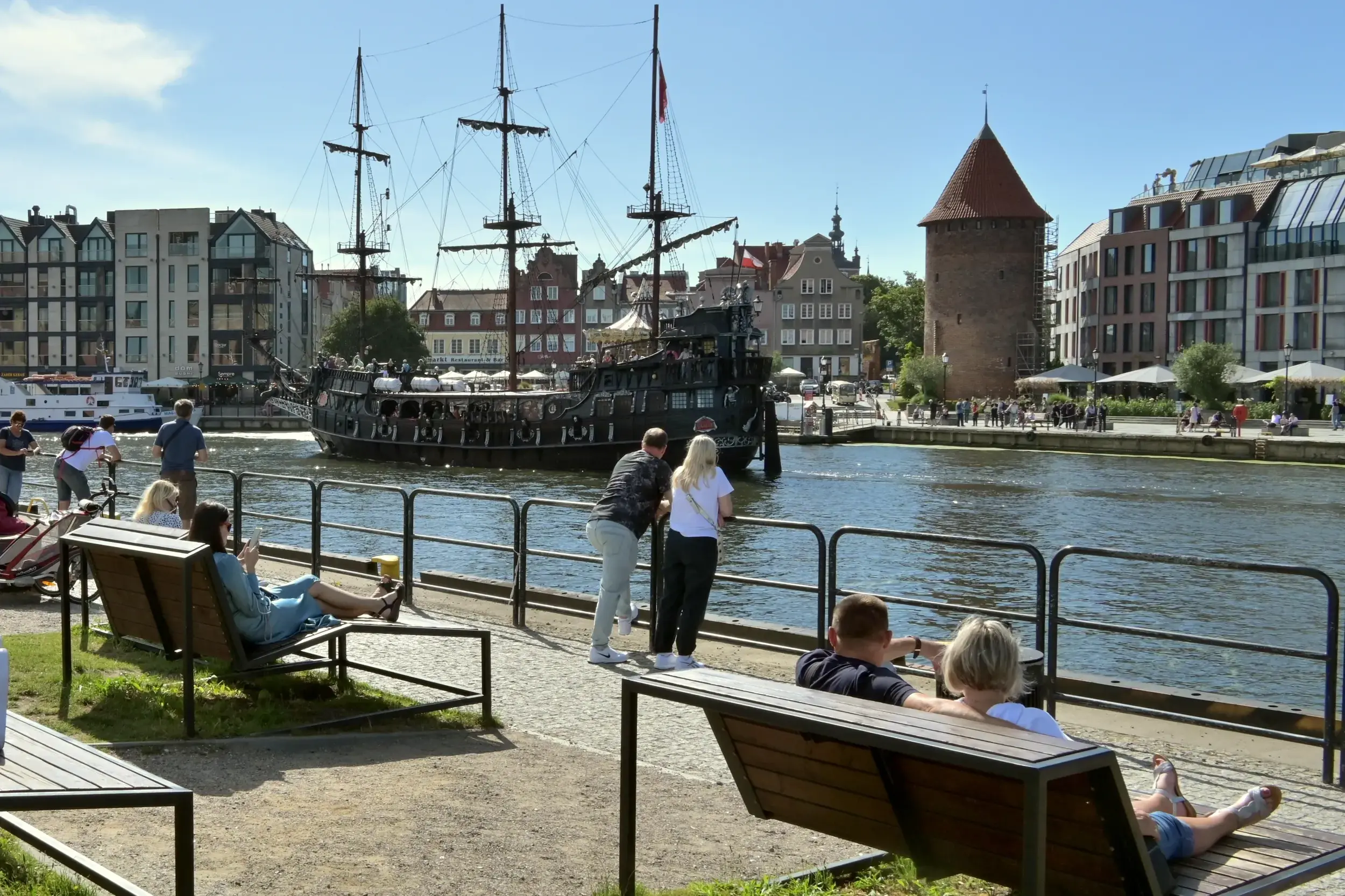 Menschen entspannen an der Uferpromenade der Mottlau in Danzig und genießen bei Sonnenschein den Blick auf ein historisches Schiff.