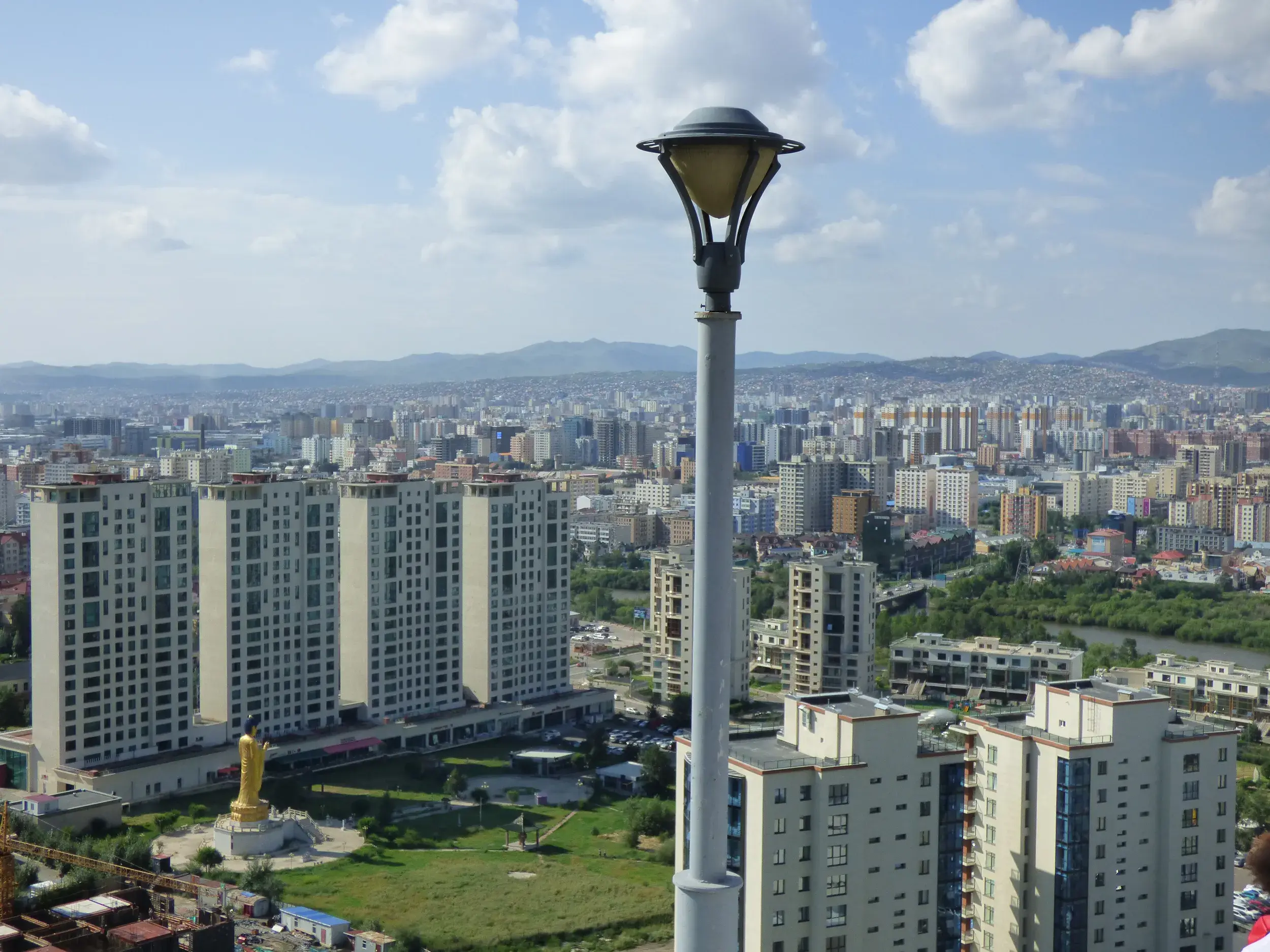 Weite Aussicht vom Zaisan-Hügel über die Hauptstadt Ulaanbaatar mit moderner Skyline und goldener Buddha-Statue am Stadtrand.