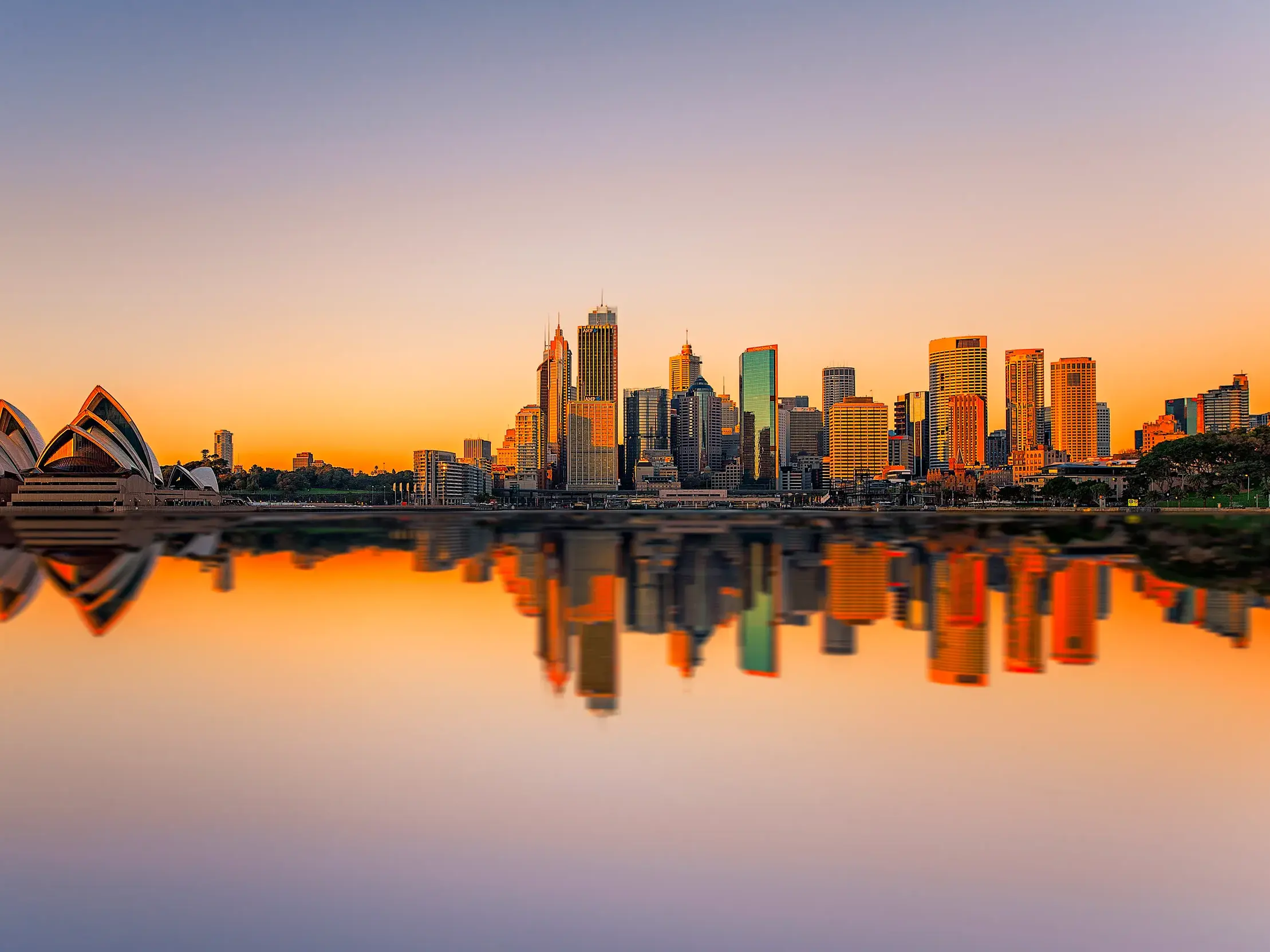 Ikonische Skyline von Sydney mit Opernhaus bei Sonnenaufgang, das die Gebäude und ihre Spiegelung im ruhigen Wasser golden färbt.