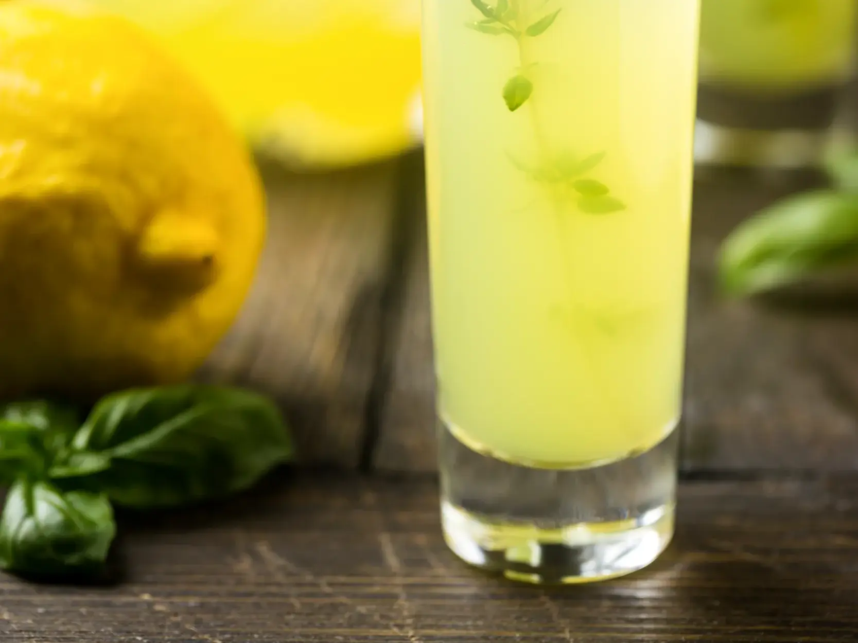 Ein Glas Limoncello, der typische Zitronenlikör der Amalfiküste, steht mit einer frischen Zitrone und Basilikum auf einem Holztisch.