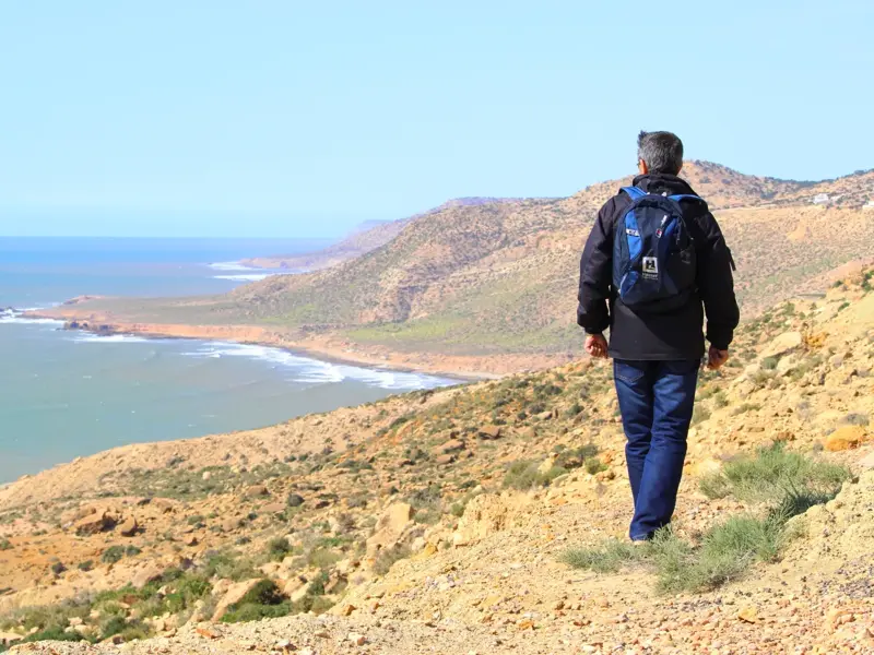 Mann mit Rucksack beim Küsten-Trekking in Marokko mit weitem Blick auf die hügelige Atlantikküste.