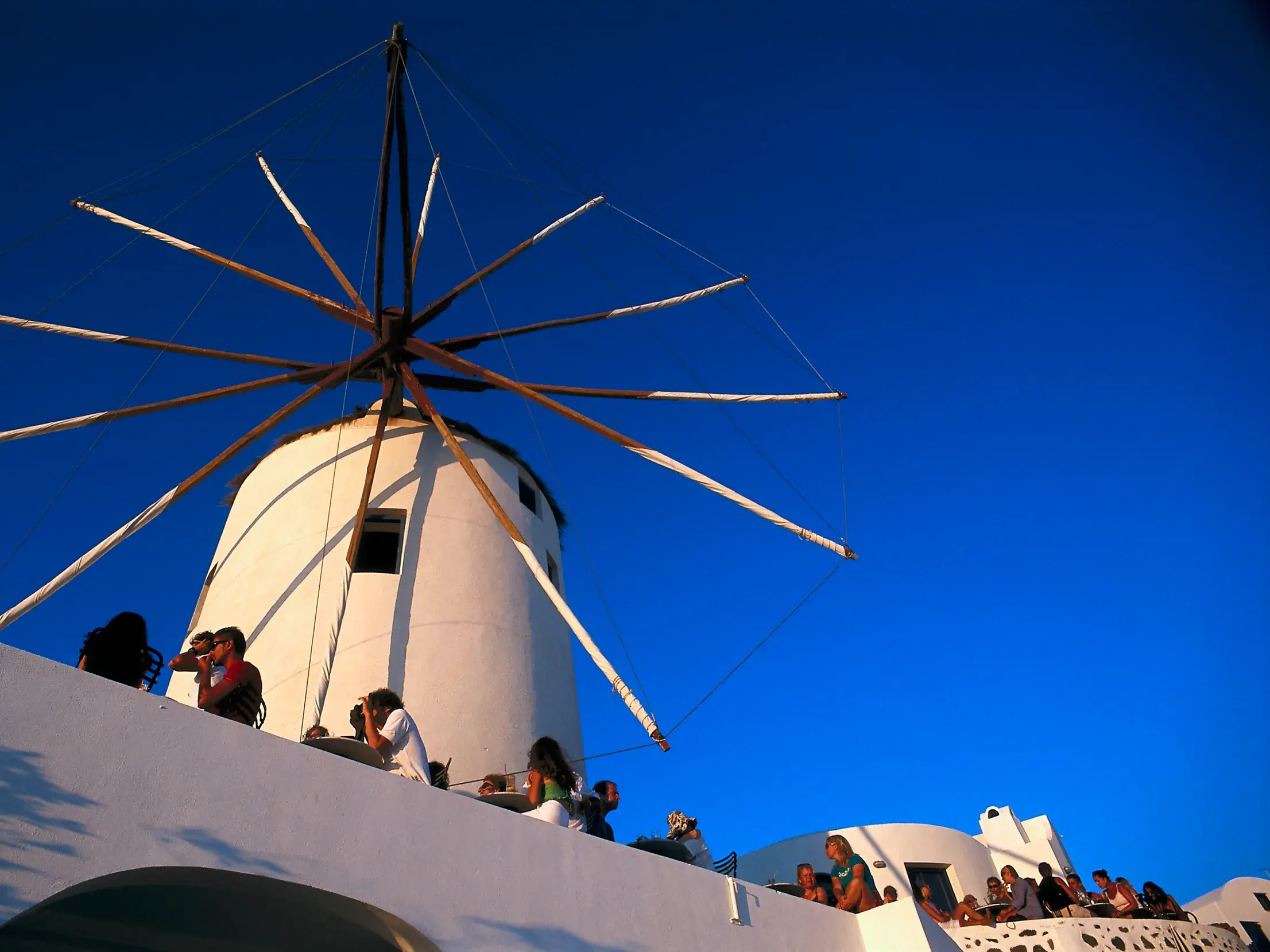 Gäste genießen die Abendstimmung auf einer Terrasse vor einer weißen Windmühle auf der Kykladeninsel Santorin.