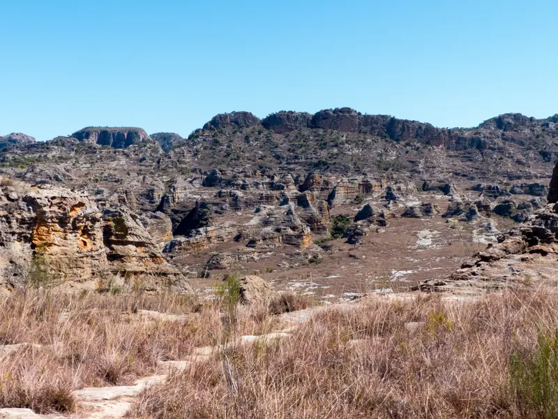 Ein Wanderpfad im Vordergrund führt durch die trockene, zerklüftete Landschaft des Isalo-Nationalparks in Madagaskar.