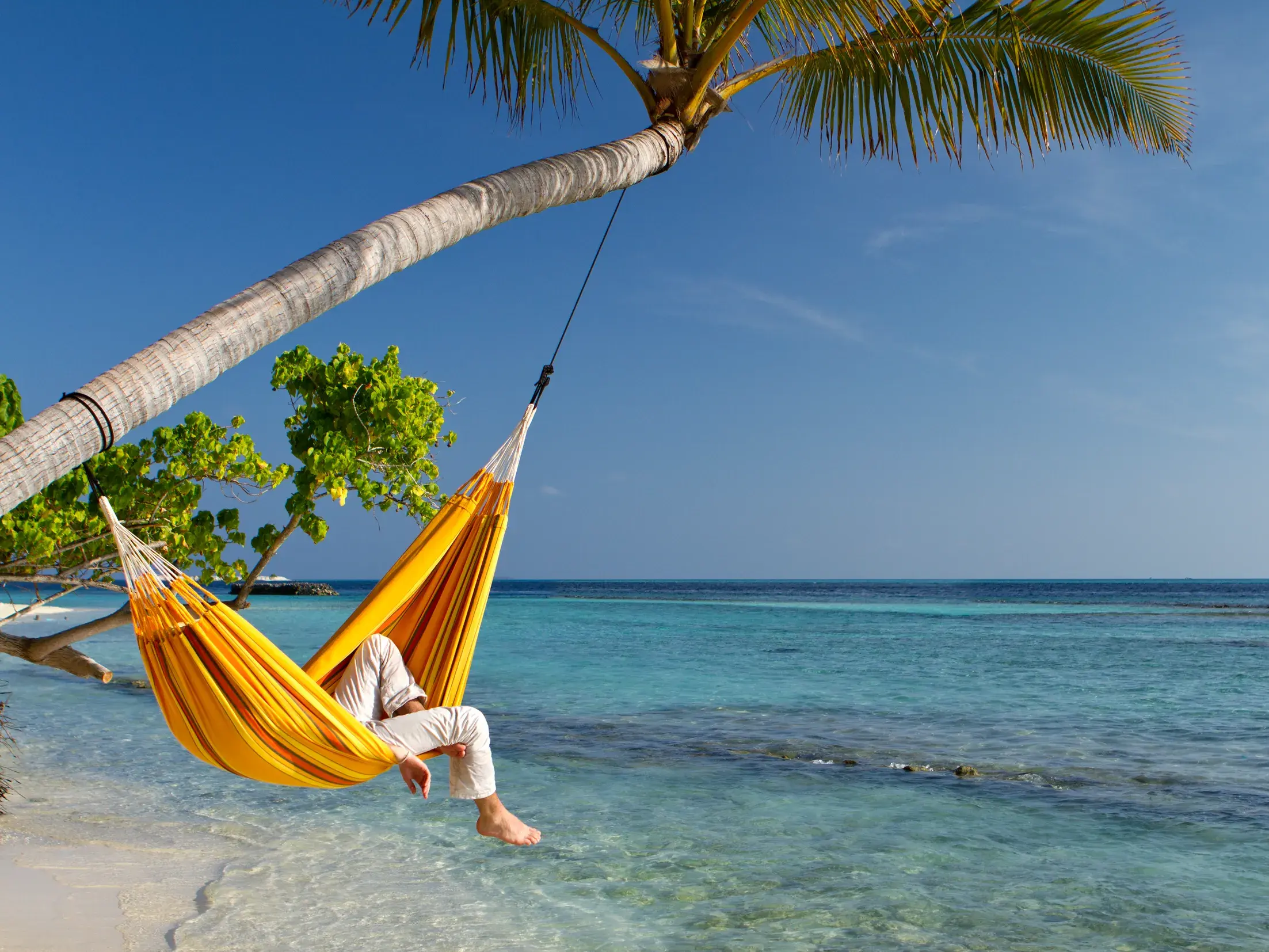 Eine Person entspannt in einer gelben Hängematte unter einer Palme am weißen Sandstrand von Samoa mit Blick auf das türkisfarbene Meer.