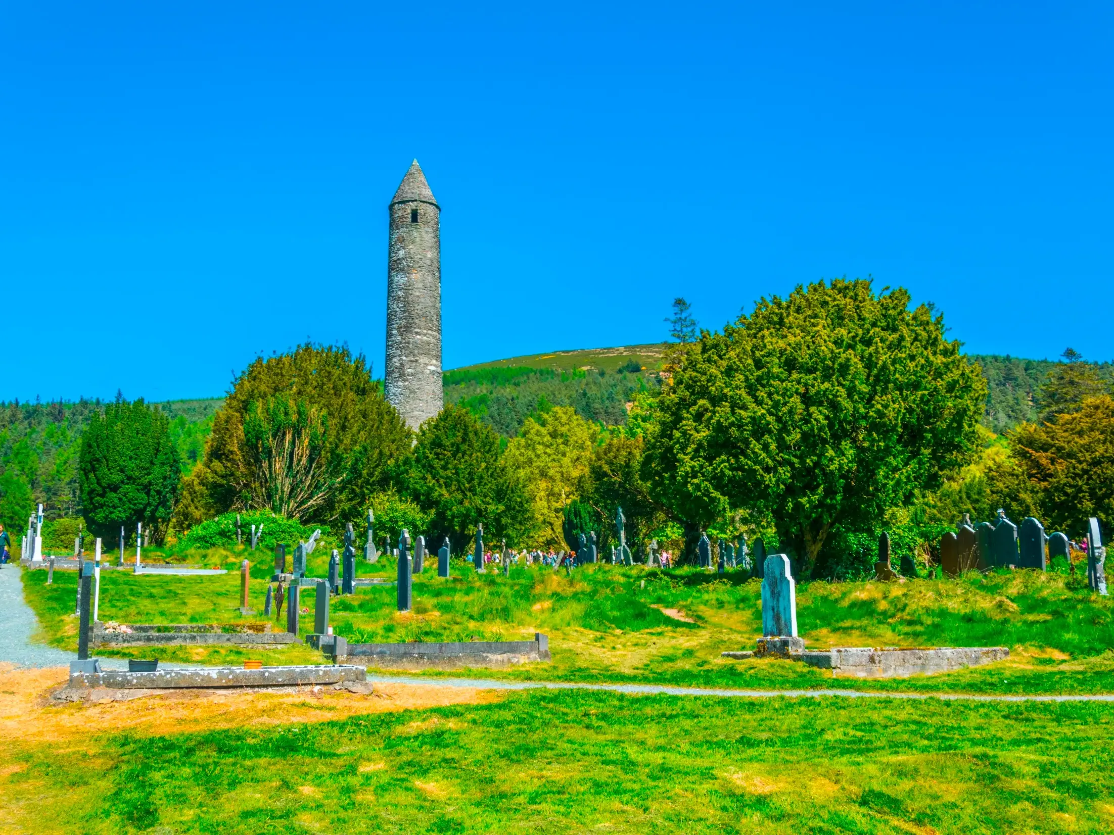 Der historische Rundturm des Wallfahrtsortes Glendalough ragt über einem Friedhof in die Höhe, umgeben von Irlands grünen Hügeln.