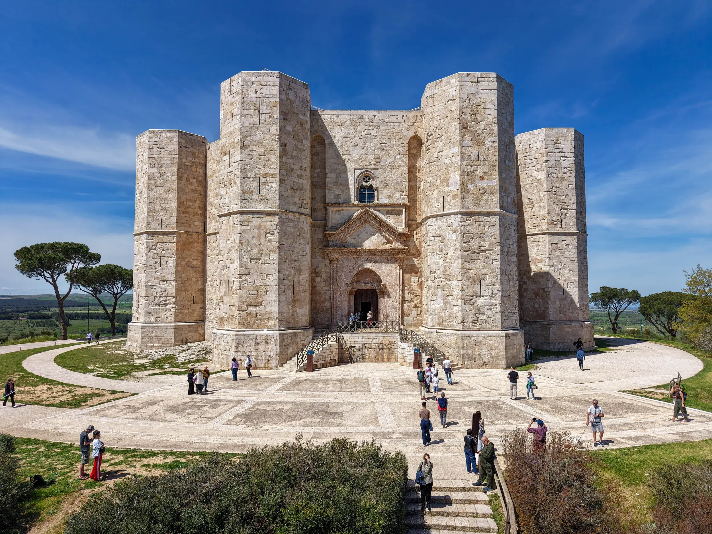 Das imposante, achteckige Castel del Monte in Apulien unter blauem Himmel. Besucher erkunden den Vorplatz des Stauferkastells.