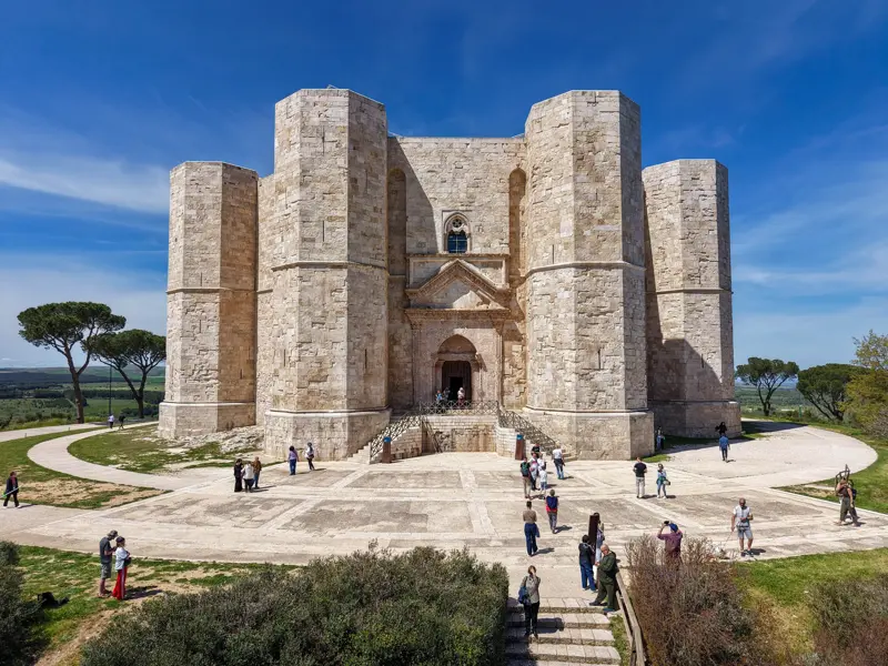 Das imposante, achteckige Castel del Monte in Apulien unter blauem Himmel. Besucher erkunden den Vorplatz des Stauferkastells.
