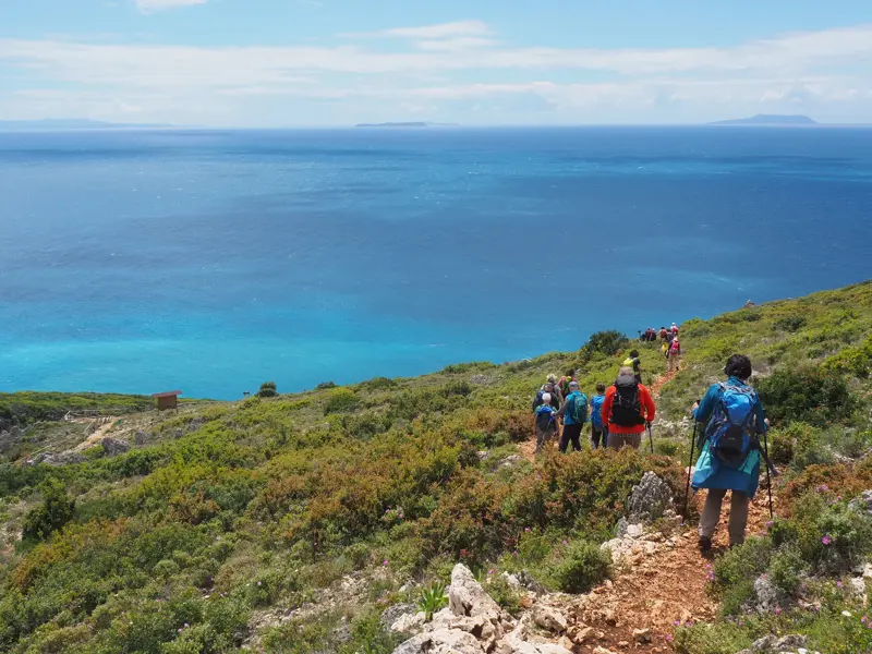 Eine Wandergruppe beim Trekking auf einem Küstenpfad an der Albanischen Riviera mit weitem Blick über das tiefblaue Ionische Meer.