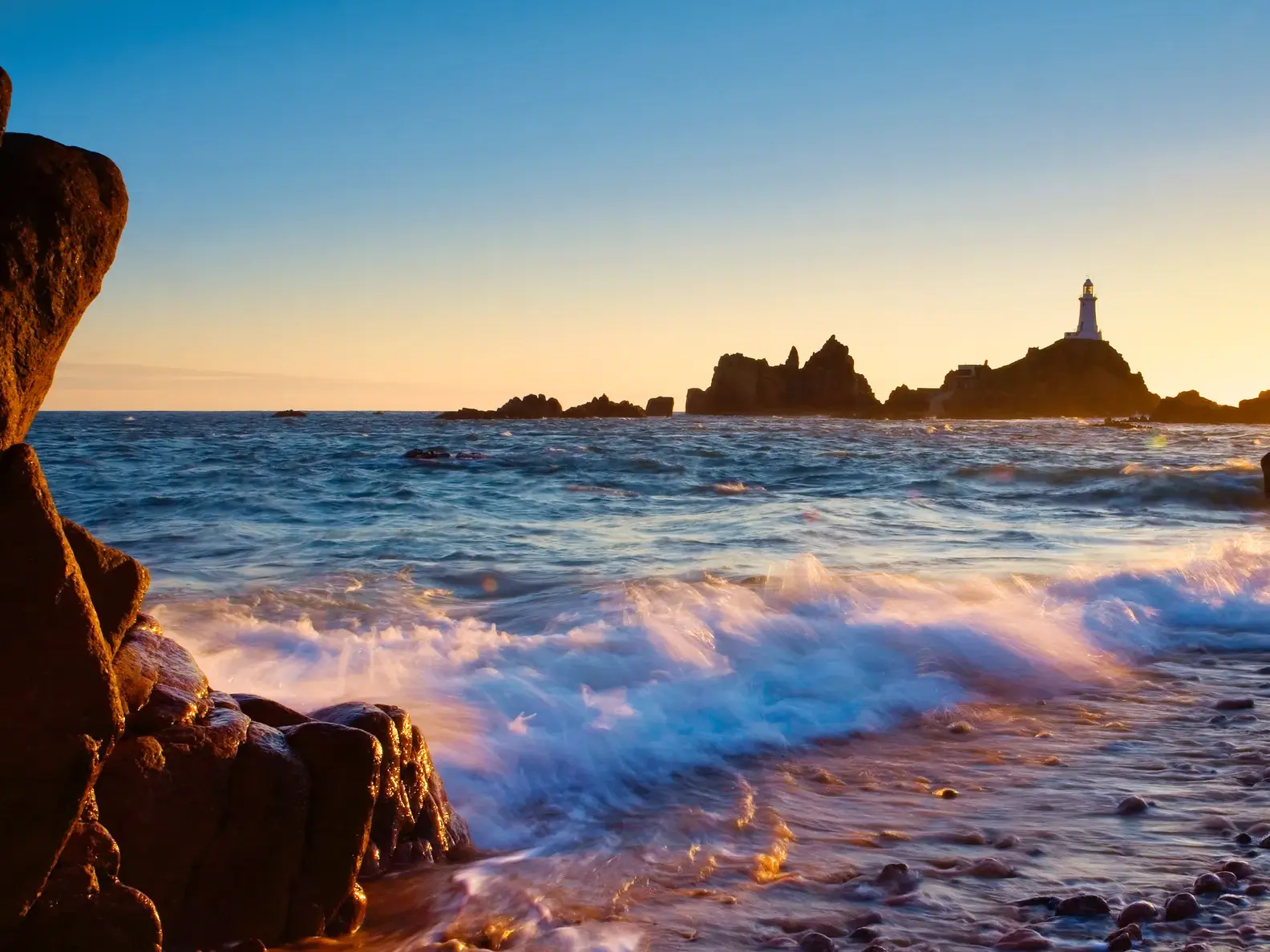 Der weiße Corbière-Leuchtturm auf Jersey, umgeben von Felsen und Wellen im warmen Licht des Sonnenuntergangs.