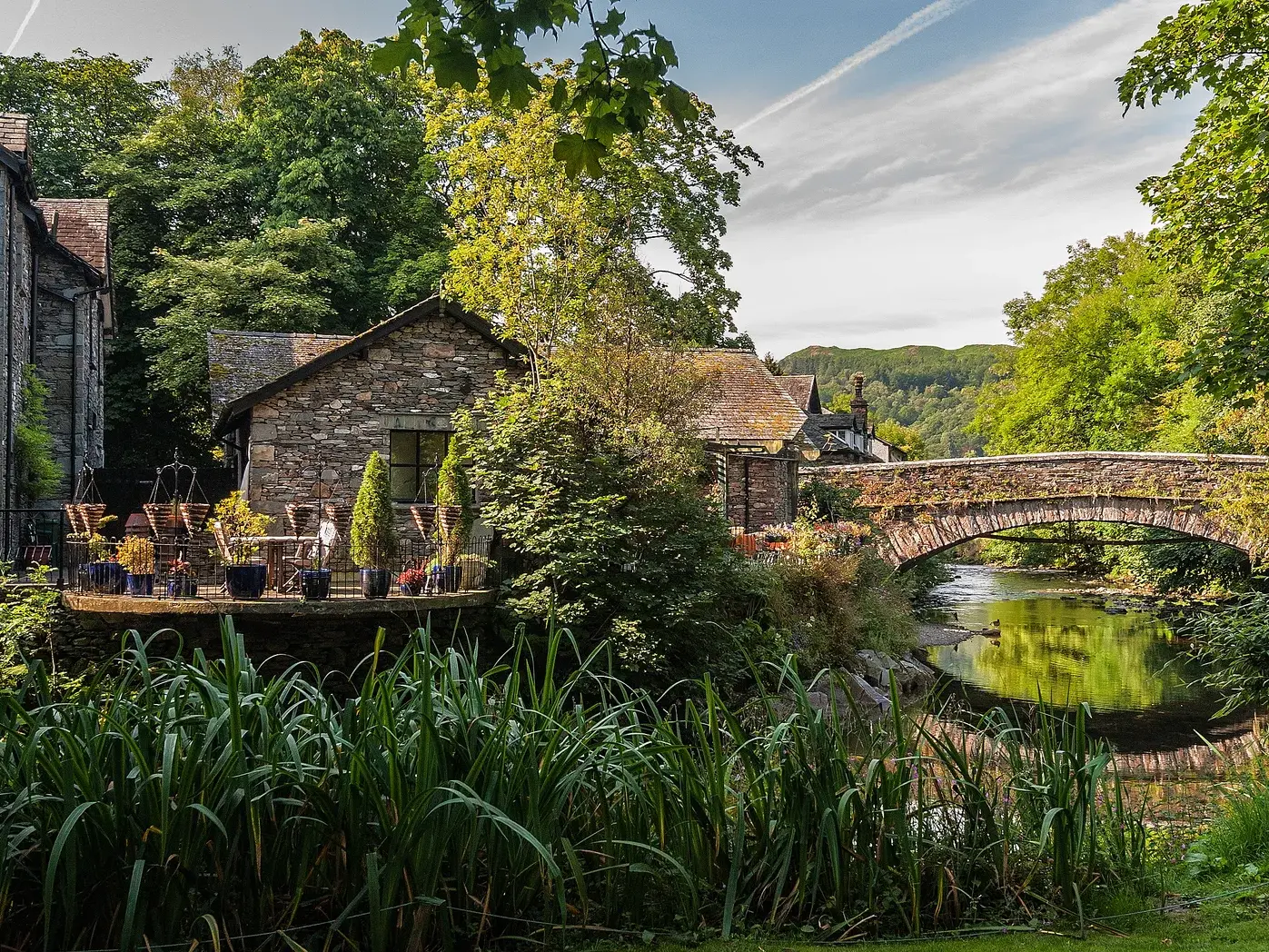 Idyllische Szene in Grasmere: ein traditionelles Steinhaus mit Terrasse am Fluss neben einer alten Steinbogenbrücke.