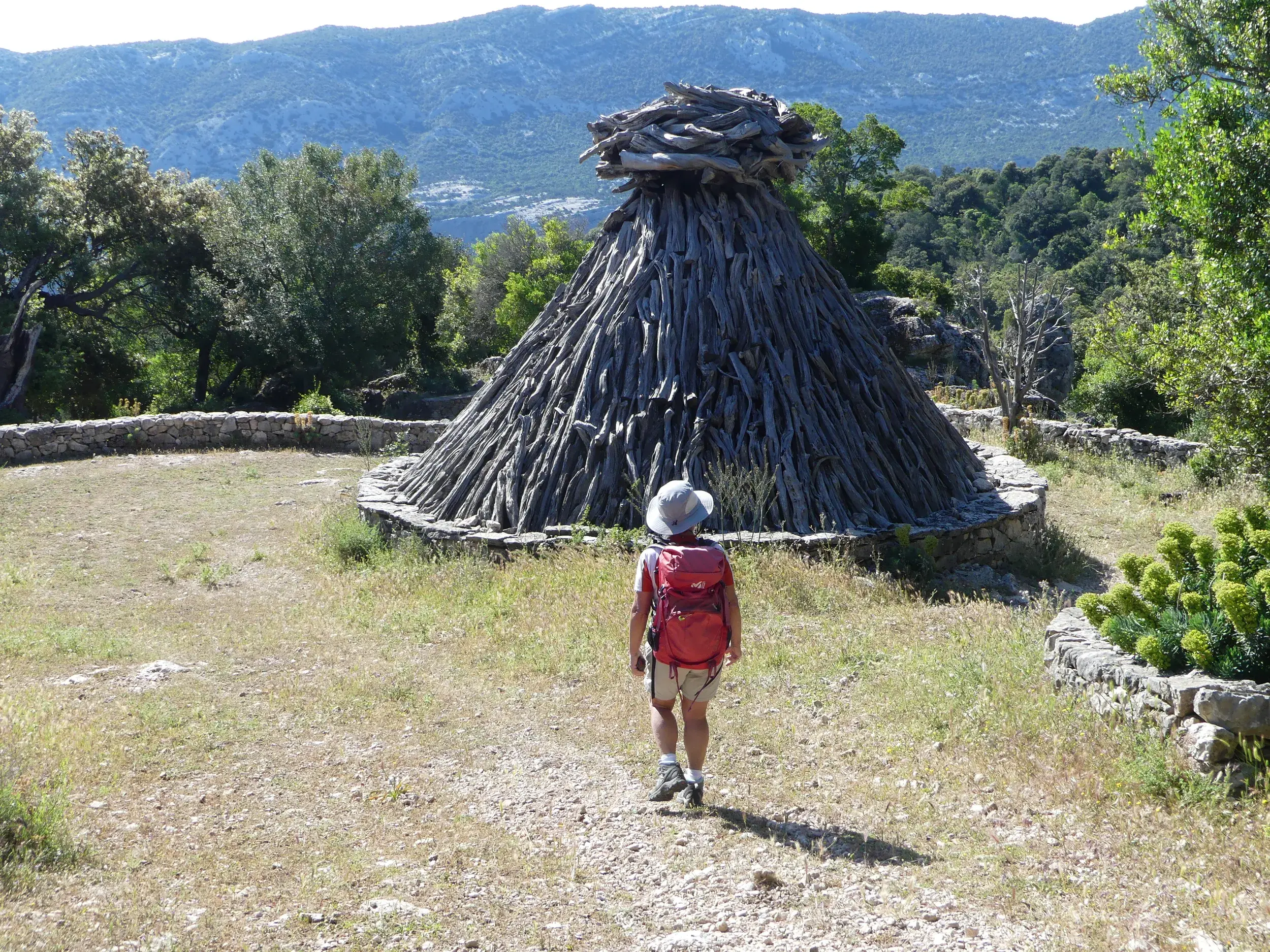 Eine Person mit Wanderrucksack nähert sich einer traditionellen Hirtenhütte aus Holz vor der Bergkulisse des Supramonte.
